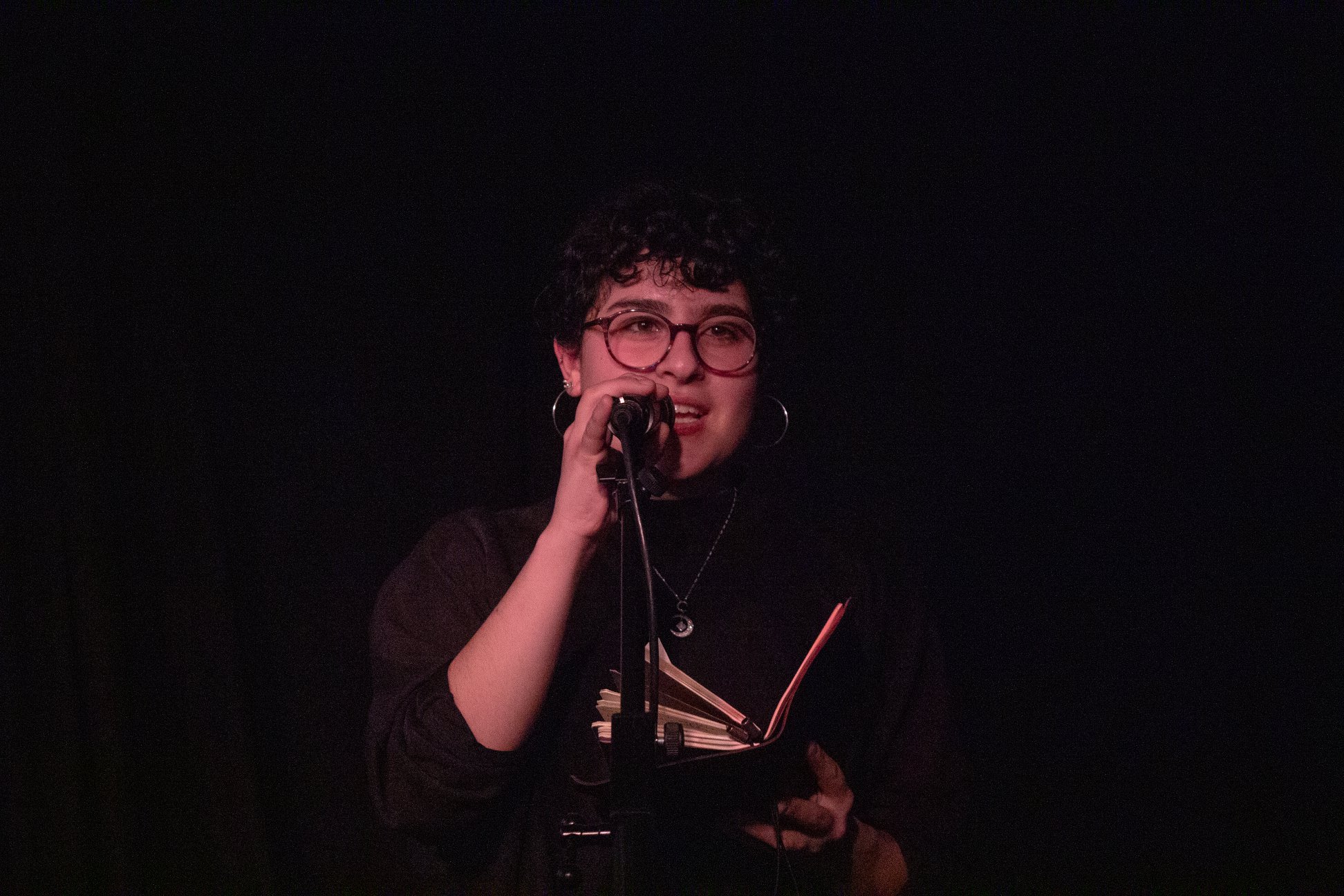A photo of writer Mahta Riazi, standing in a dimly-lit room, holding a microphone and reading from a book. She has curly bangs and wears large round glasses.