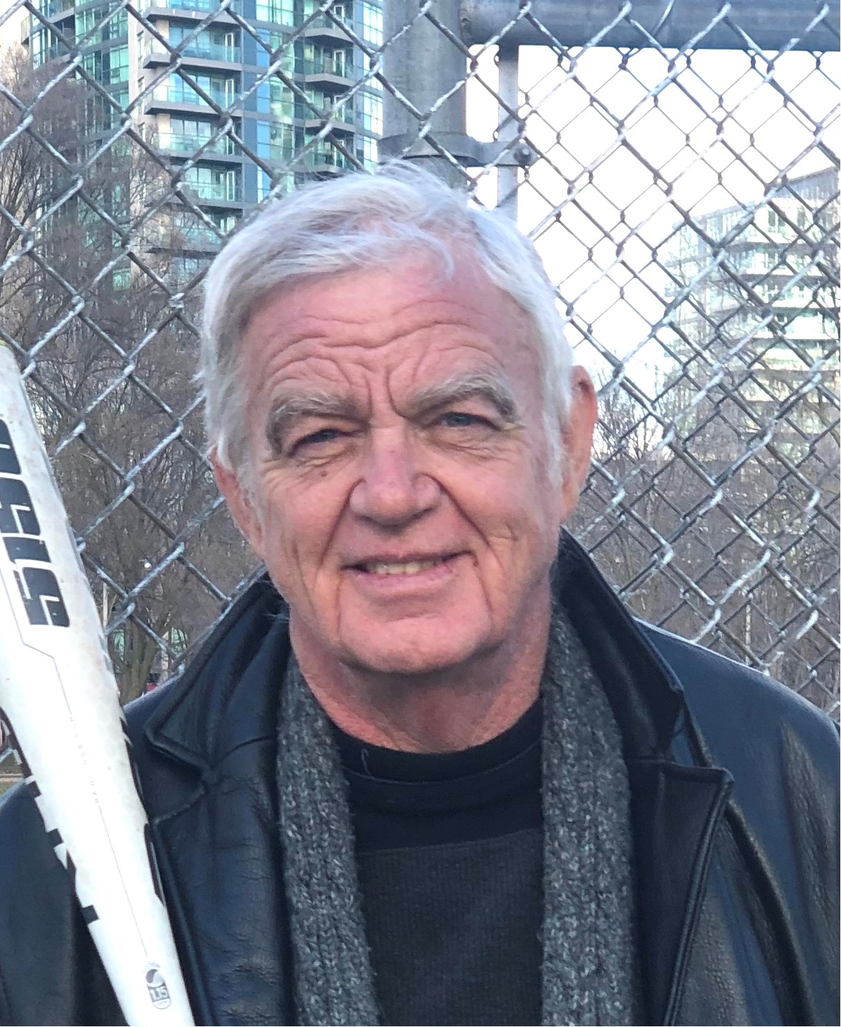 A photo of writer Peter Unwin. He is a light skin-toned man with short grey hair, wearing a leather jacket over a dark wool sweater. He holds a metal baseball bat and is standing against the chain link fence of a baseball diamond, with a condo building in the background.