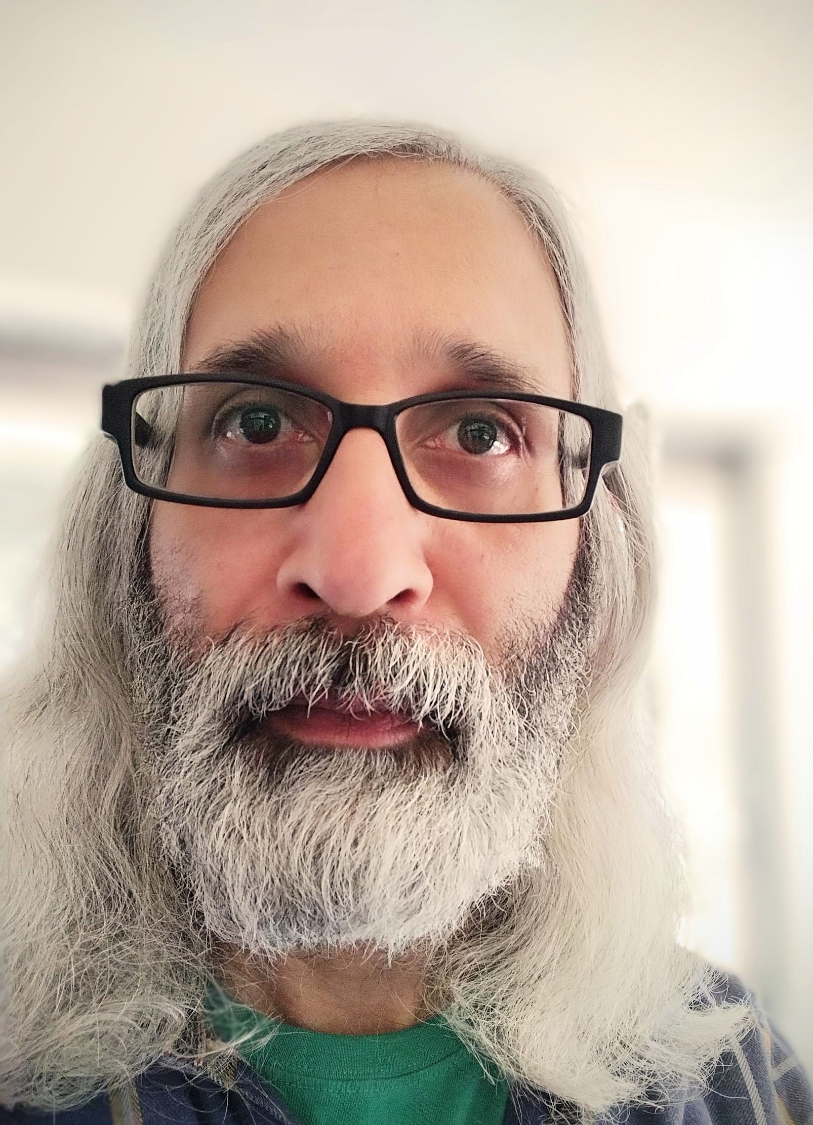 A close-up photo of Ayaz Pirani, a man with long grey hair and a beard. He is wearing black-rimmed glasses and looking into the camera.