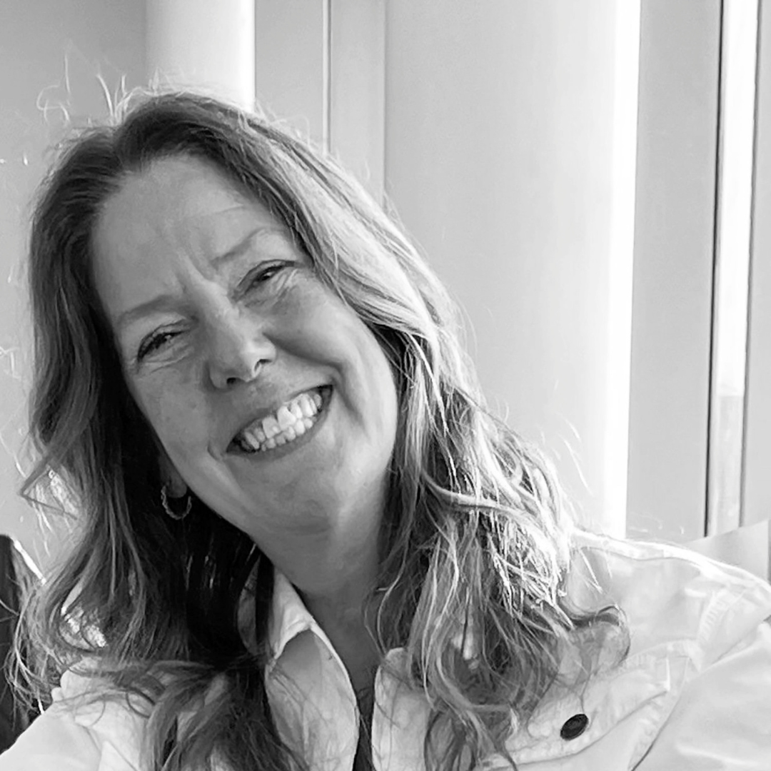 A black-and-white photo of writer Louise Ells. She is a light skin-toned woman with medium length light hair, leaning over to one side with a huge smile on her face.