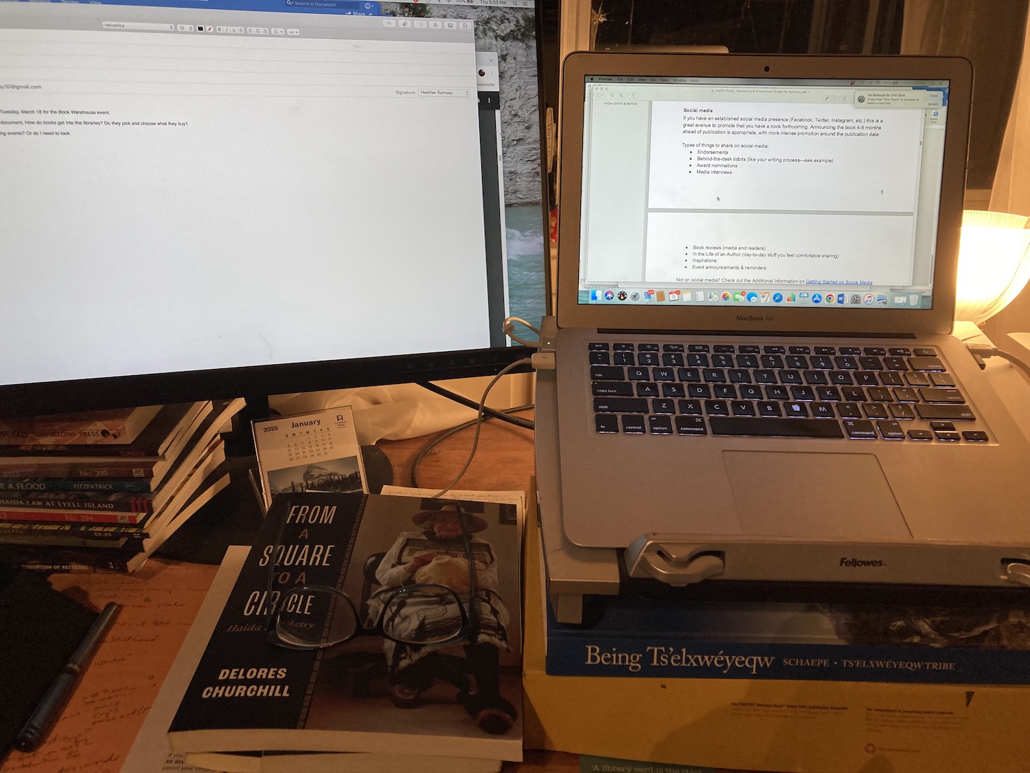 An open laptop next to a secondary monitor. A collection of Haida Gwai related books are stacked on the work surface.
