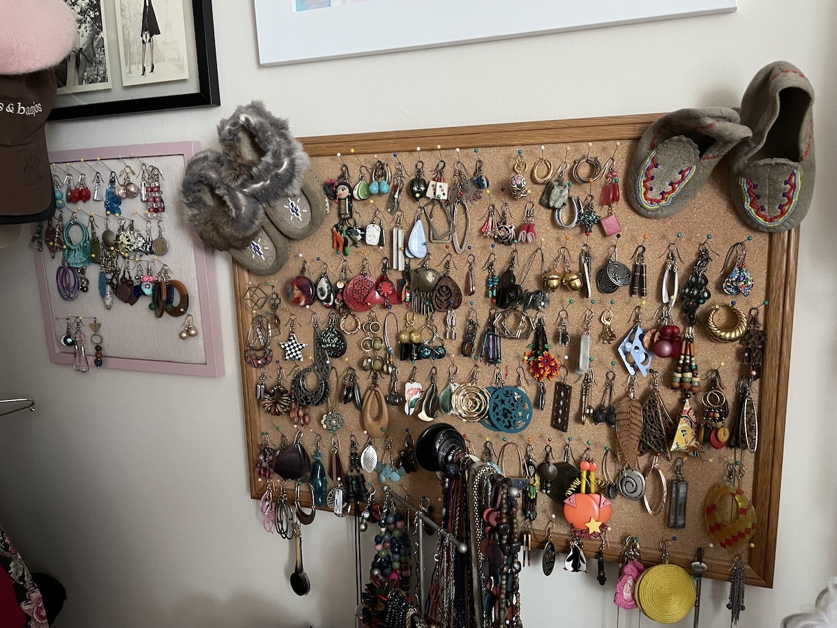 Dozens of pairs of earrings hang on two corkboards on the wall, in a variety of colours, shapes, and metals. Two mini moccasin pairs sit at the upper corners of the larger corkboard. The collection is lively and funky.