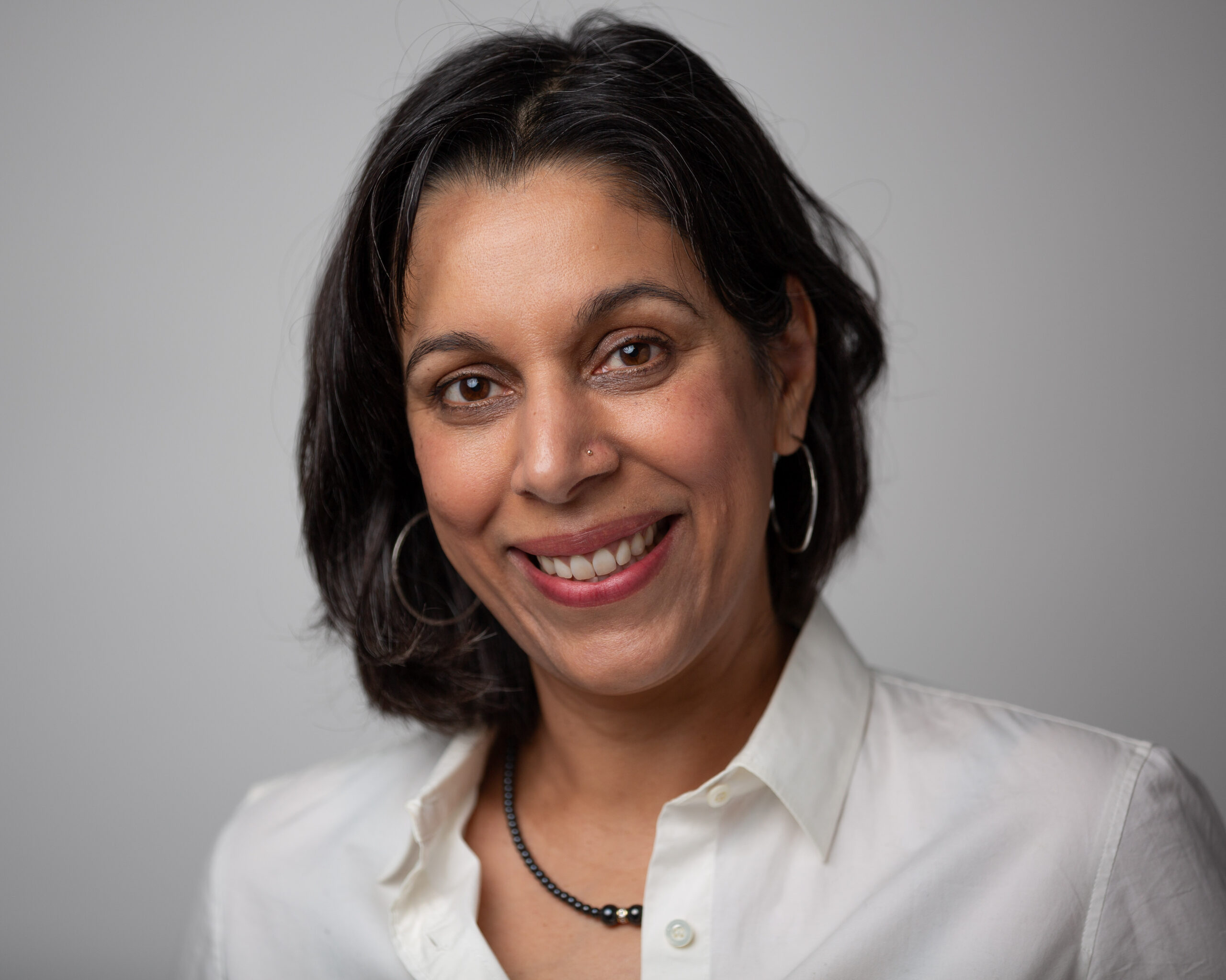 A photo of author Kirti Bhadresa. She is a brown-skinned woman with short dark hair. She is wearing a white-collared shirt and necklace and smiling into the camera. Photo credit Jeremy Fox.
