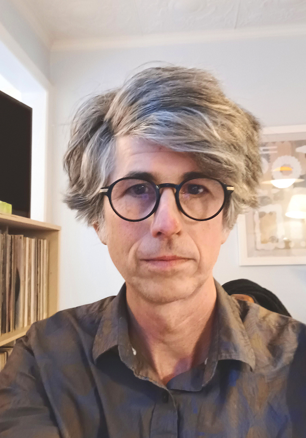 A photo of writer and book designer Malcolm Sutton. He is a light skin toned man with voluminous straight grey hair, wearing black round glasses and a grey button up shirt. He stands next to a shelf of vinyl records.