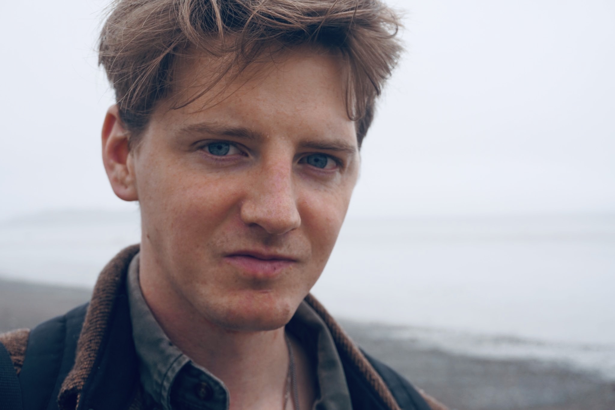 A photo of Luke Francis Beirne. He is a light-skin toned man with short light brown hair and blue eyes. He is standing on a beach in front of a body of water and looking into the camera. 