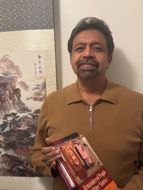 A photo of writer Pratap Reddy. He is an Indian man with fluffy dark grey hair and a trim goatee, wearing a brown zip sweater. He smiles, holding copies of his books with Remaindered People & Other Stories at the top of the stack.