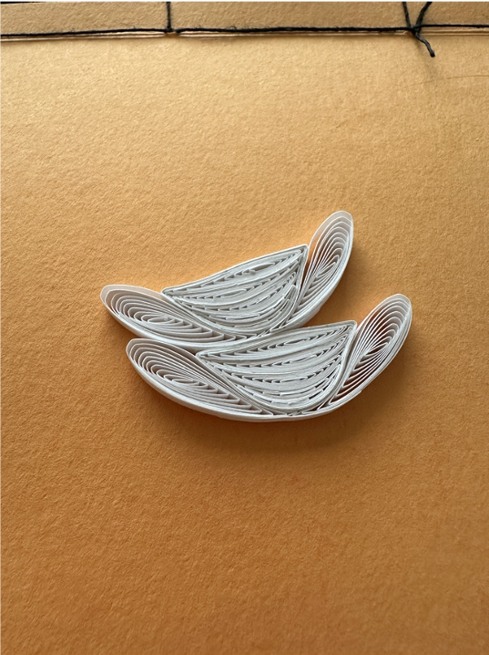 Two quilled paper shapes that curve and fold into each other, resembling the vertebrae of a human spine, lay on a light brown surface.