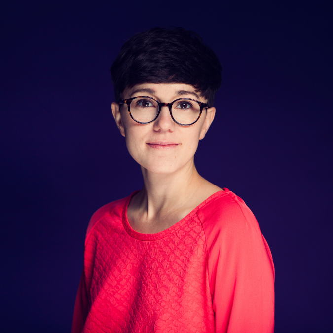 A photo of playwright Sanita Fejzic. She is a light skin-toned woman with short, bowl-cut dark hair and dark framed glasses. She wears a bright pink shirt and stands before a dark blue background.