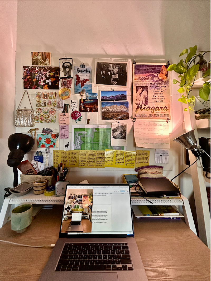 A photo of Joni Murphy's workspace. An open laptop on a desk sits before a corkboard pinned with photos, postit notes, a glittery purse. Two lights up-light the contents of the corkboard. The vine of a houseplant shows in the right corner.