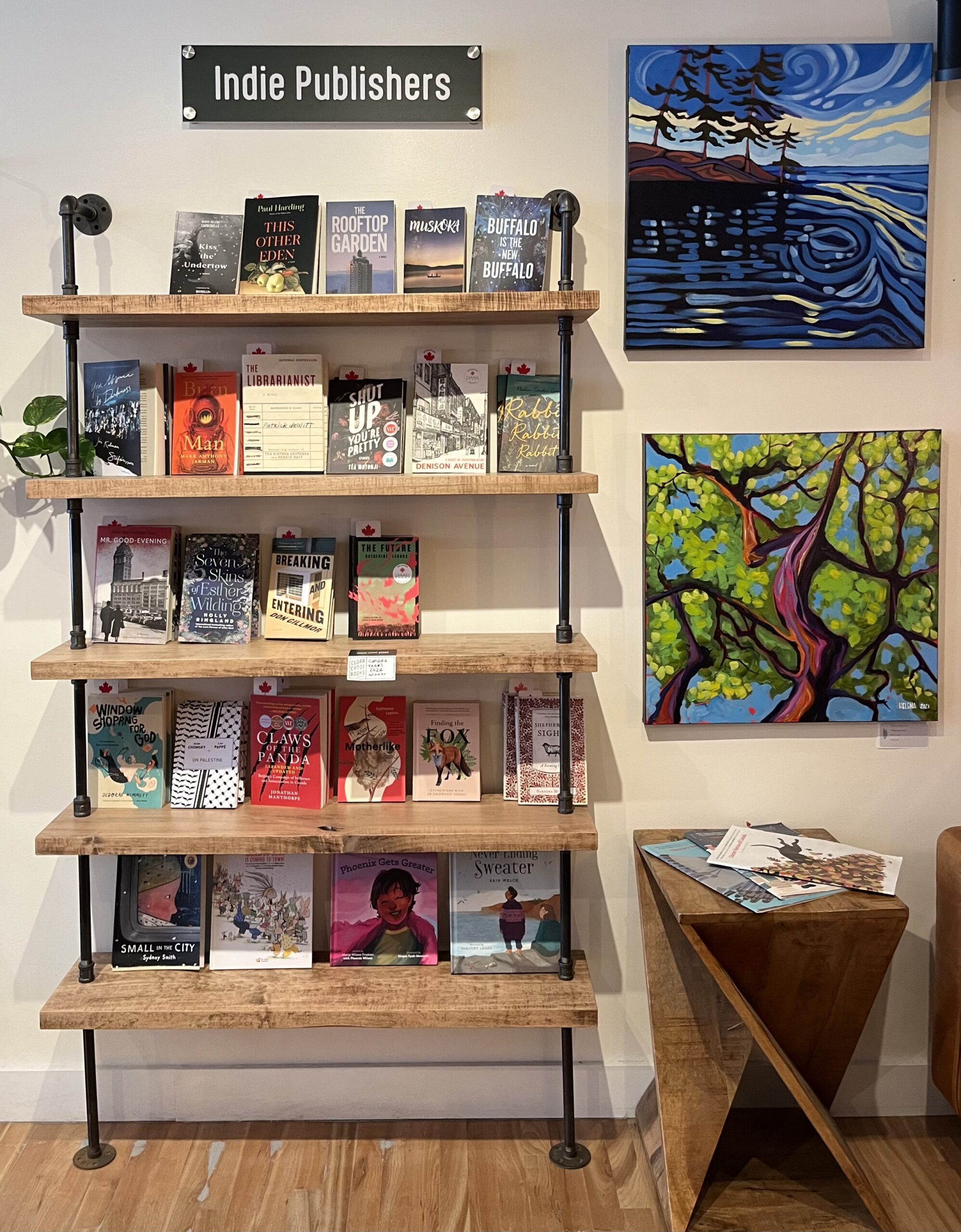 A photo inside Cedar Canoe Books in Huntsville, Ontario. The photo shows a wooden bookshelf secured to a wall with black steel pipes. There are several books displayed on the shelf with a sign above reading Indie Publishers. There are two paintings of nature scenes on the wall beside the shelf. 