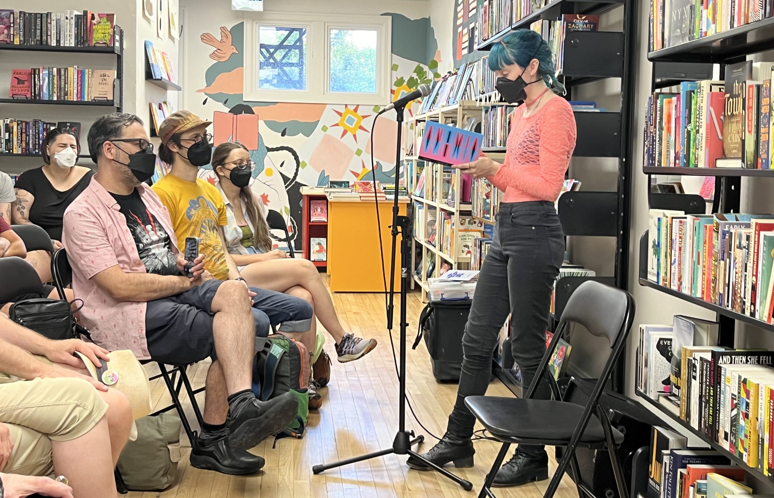 A photo of author Jade Wallace reading from their book Anomia at Take Cover Books in Peterborough, Ontario for a seated audience. There are bookshelves all around the room. 