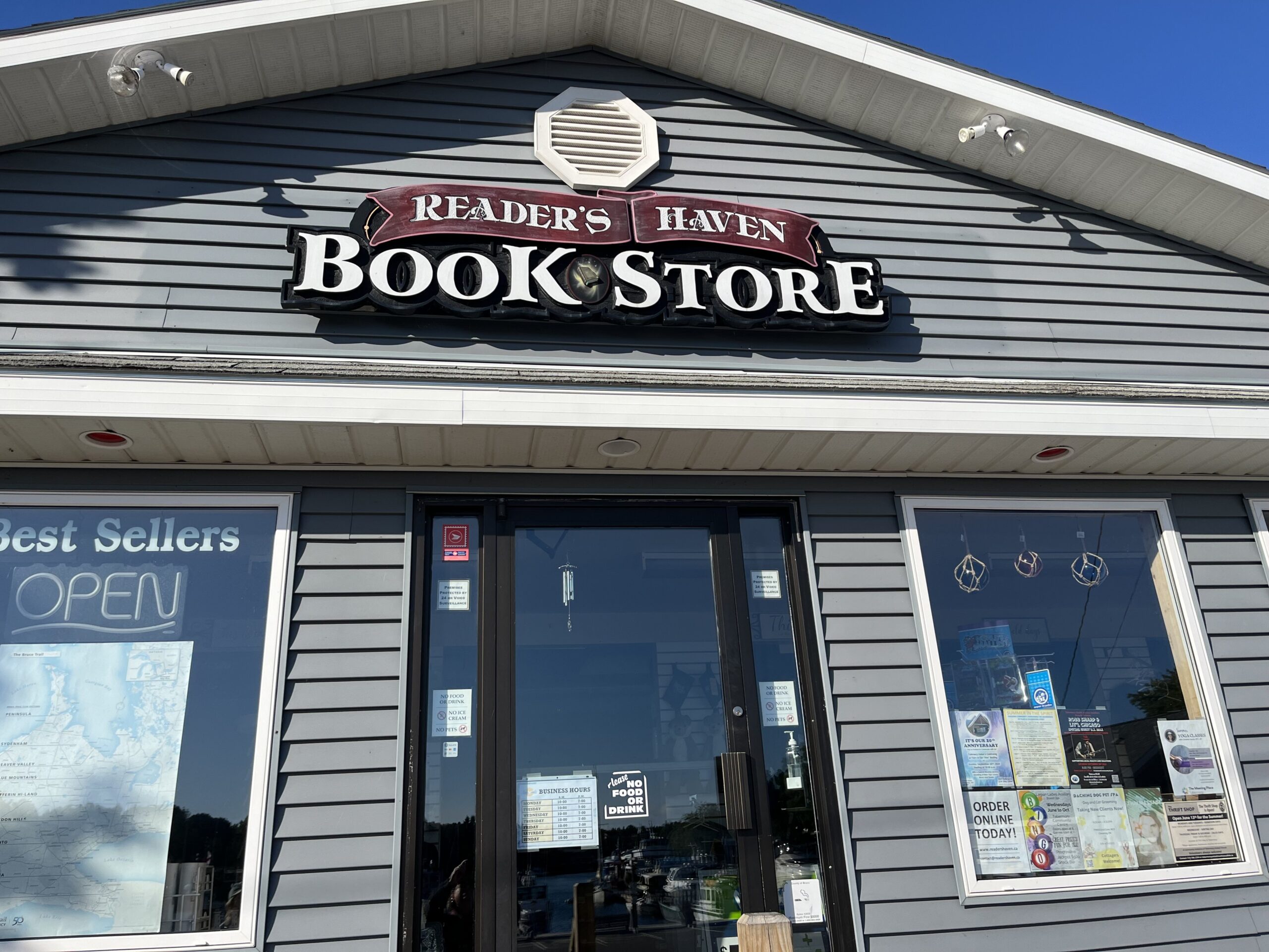 The storefront at Reader's Haven Bookstore in Tobermory, Ontario. It is a house with grey vinyl siding and two windows on either side of a glass entrance door. There are books displayed in one of the windows and a map in the other. 