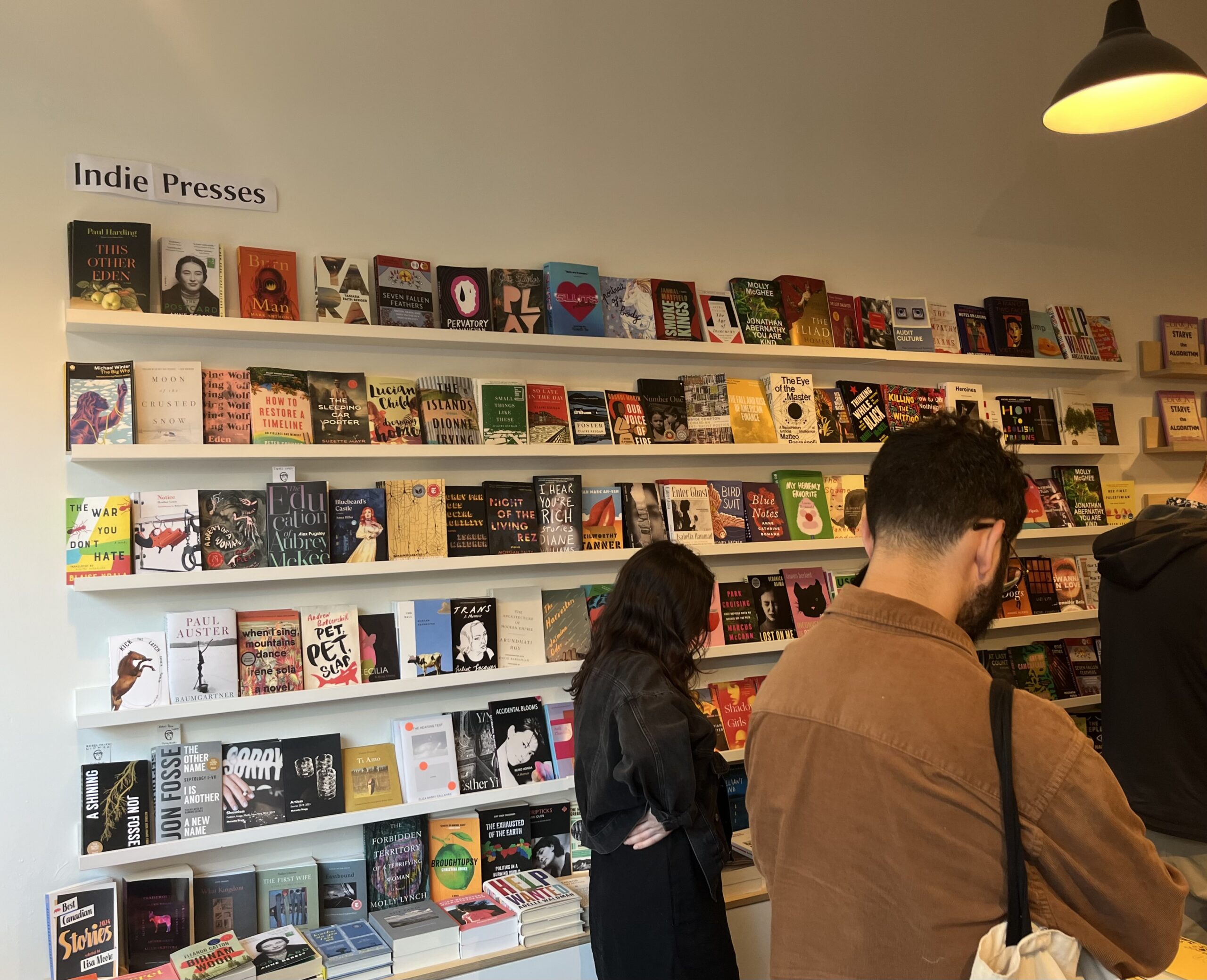 A photo of the interior space at Flying Books in Toronto, Ontario. There is a wall of books with a sign above reading Indie Presses. There are a couple of people browsing. 