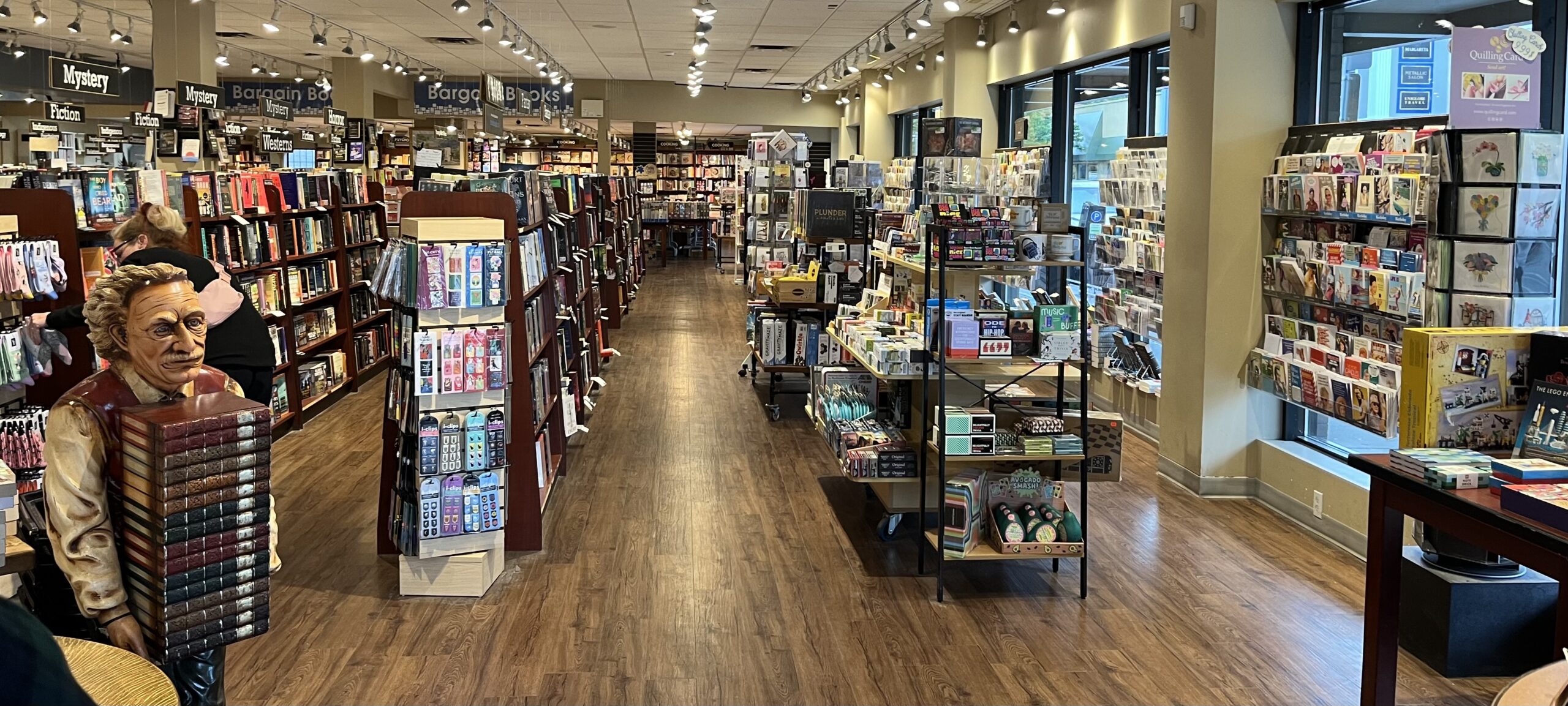 A photo from the entrance of Mosaic Books in Kelowna, BC looking into the store. The store has hardwood floors and several rows of bookshelves with books on them, as well as greeting cards on one side of the store, and a statue of a person holding a large stack of books at the front. 