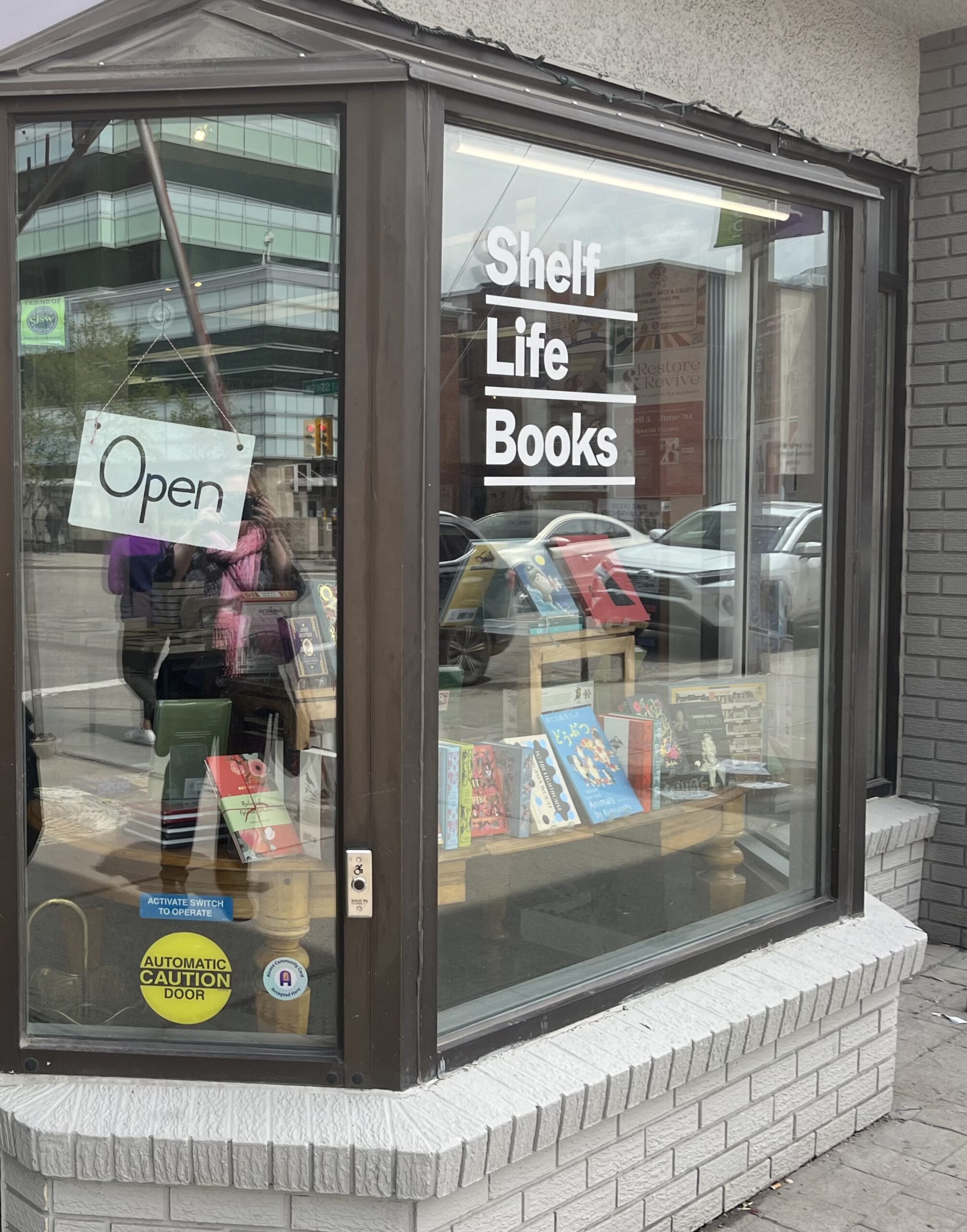 A photo of the front window of Shelf Life Books in Calgary, Alberta taken on a slight angle. There is an open sign on the window and a book display. 