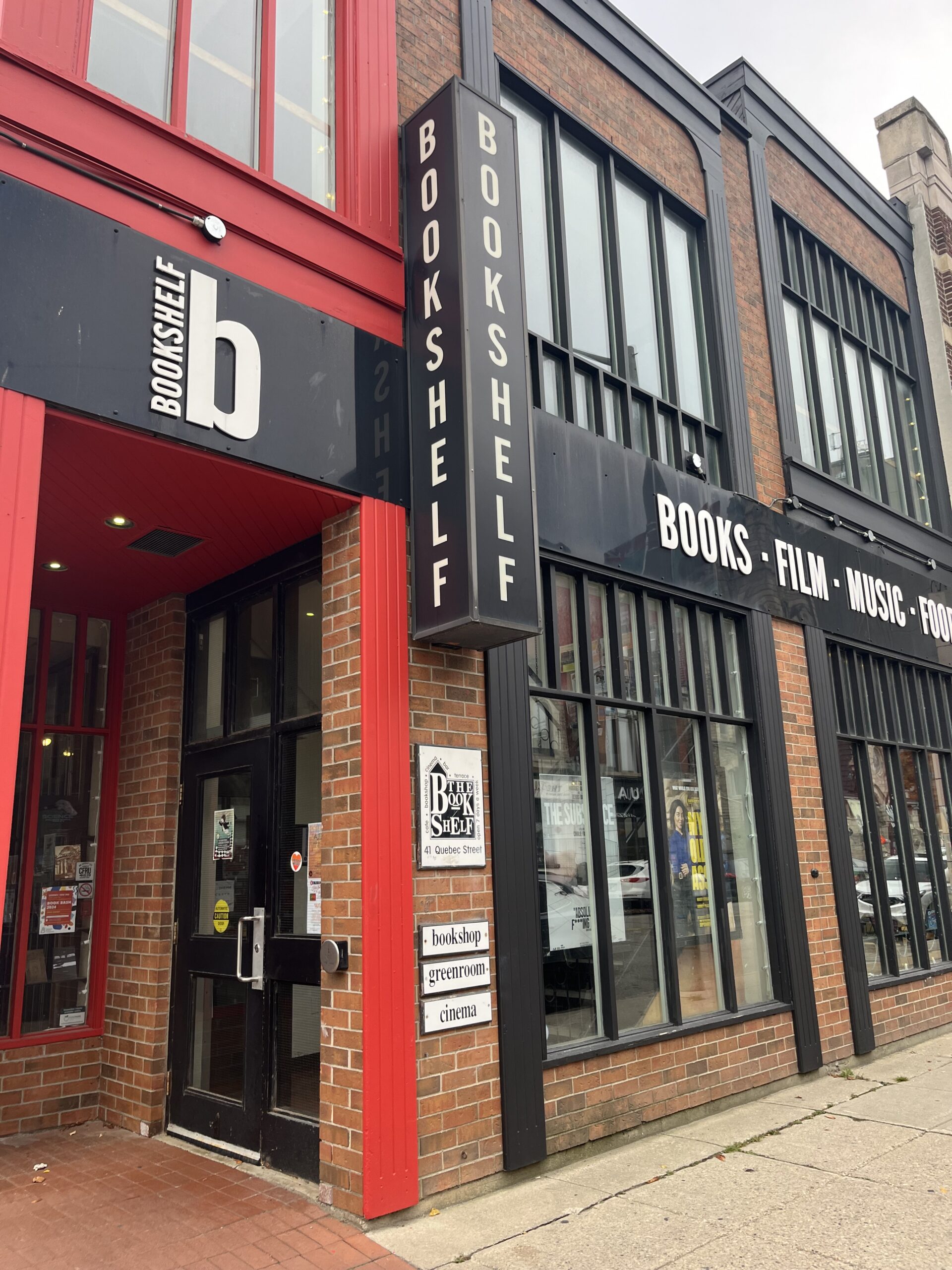 A photo of the storefront at Bookshelf in Guelph, Ontario. it is a brick building with red and black accent strips and a large sign reading "Bookshelf, Books. Film. Music. Food."