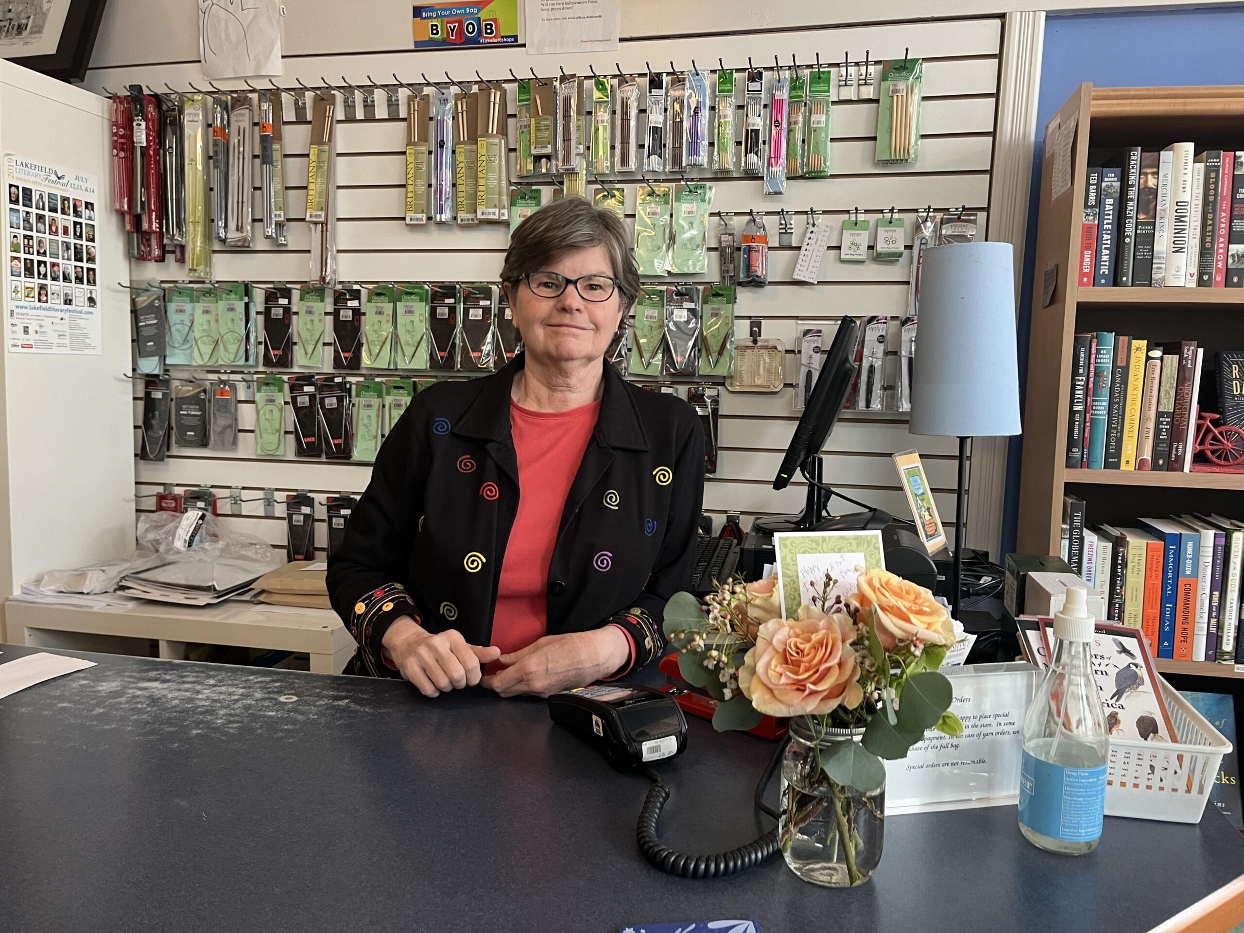 A photo of a light-skin-toned woman with short brown hair and black-rimmed glasses. She is standing behind the counter at Happenstance Books and Yarns. Behind her are an array of knitting needles and tools and a bookshelf with books. 