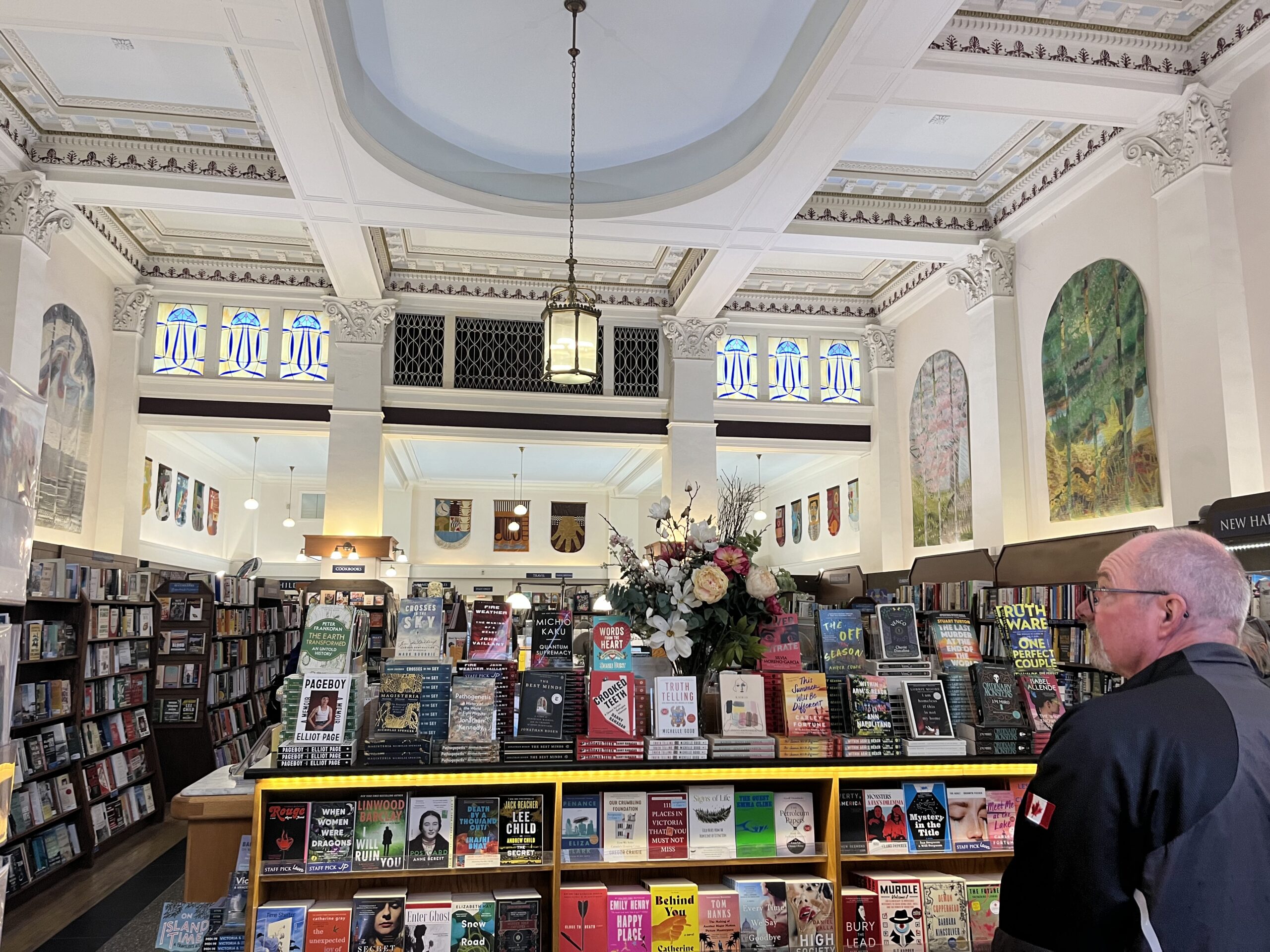 A photo of the interior of Munro's Books in Victoria, BC. The store features high coffered ceilings, stained glass, and artwork along the walls. There are bookshelves along the walls as well as down the centre of the store. 