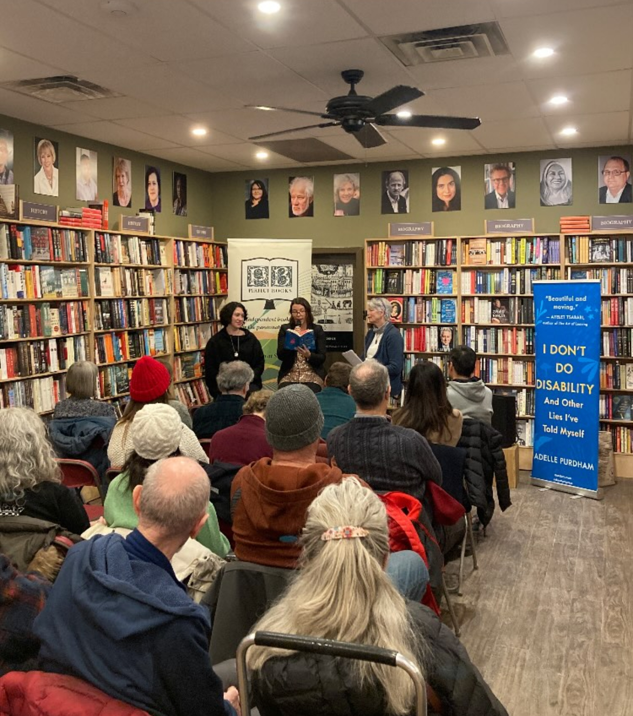A photo of a book reading at Perfect Books in Ottawa, Ontario. In the photo, three authors sit in front of a small seated audience. There is a banner sign beside where they sit reading "I Don't Do Disability and Other Lies I've Told Myself, Adele Purdham." Along the walls are bookshelves full of books. 