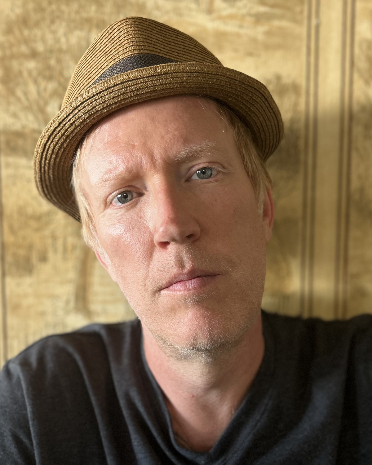 A photo of poet Scott Nolan. He is a light-skin toned man with blonde hair peeking out from under a straw fedora, wearing a dark grey tshirt.