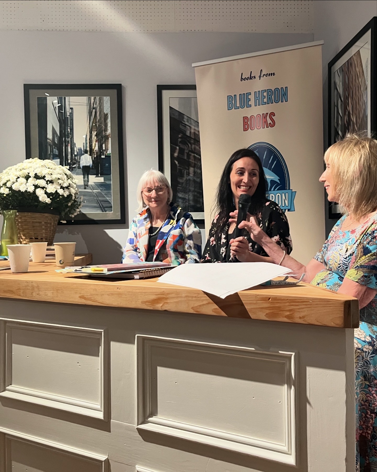 A photo taken at Blue Heron Books in Uxbridge, Ontario during a book launch. Three women sit behind a desk engaged in conversation. 