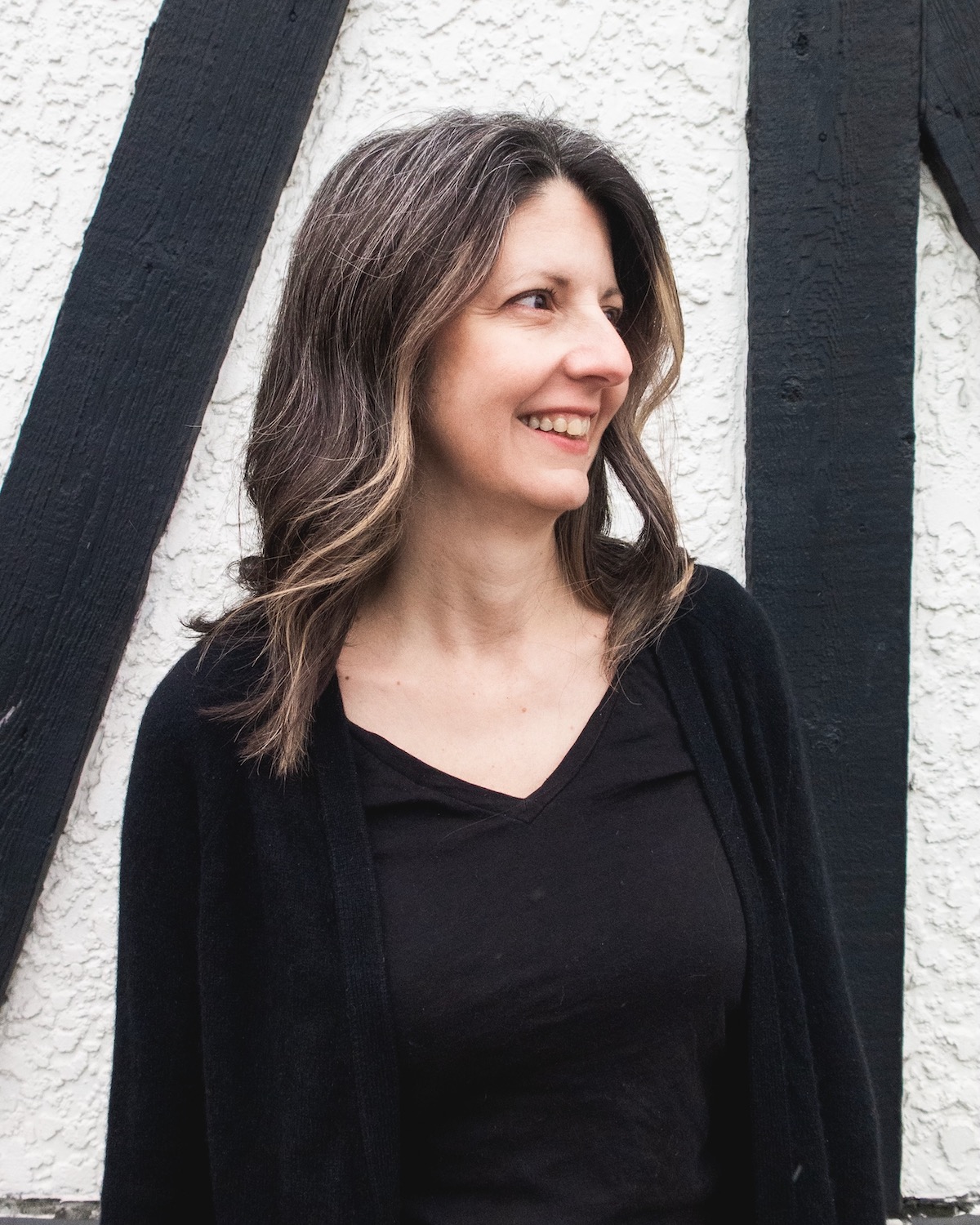 A photo of poet Holly Flauto. She is a light skin toned woman with medium length brown hair, wearing a black t-shirt and cardigan, and standing before a white stucco building with black, tudor-style wooden beams.