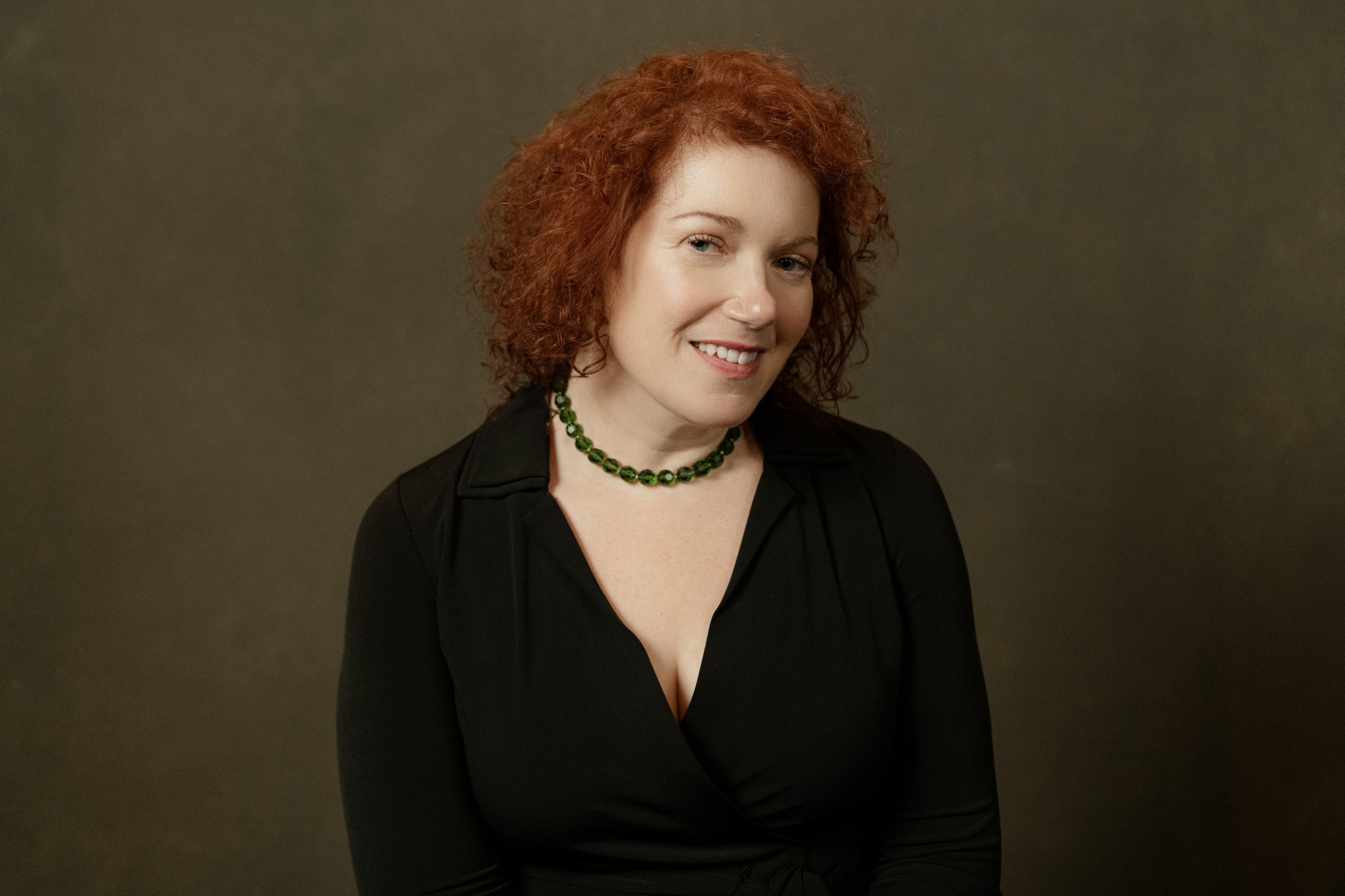 A photo of Kim Fahner. She is a light-skin-toned woman with short curly red hair. She is wearing a black long-sleeve shirt with a green beaded necklace and smiling into the camera.