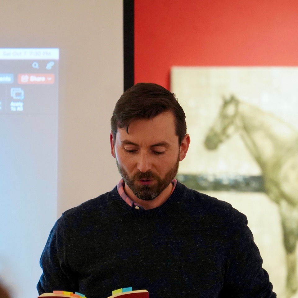 A photo of author Matthew Gwathmey. He is a light-skin-toned man with short dark brown hair and a trimmed beard. He is wearing a black sweatshirt and standing in front of a wall with a painting of a horse. He is reading from a book. 