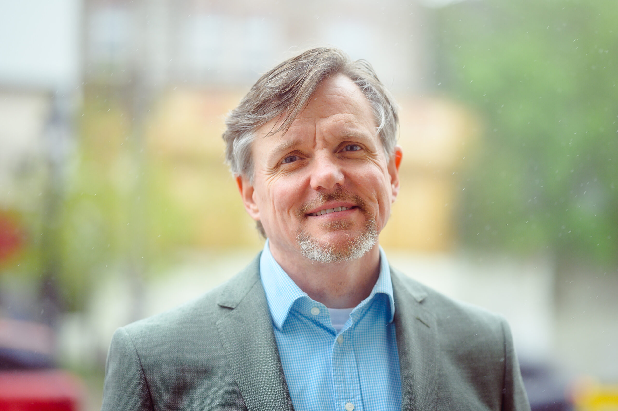 A photo of author Paul Moorehead. He is a light-skin-toned man with grey hair and short facial hair. He is wearing a blue button-up shirt under a grey suit jacket and smiles into the camera. 