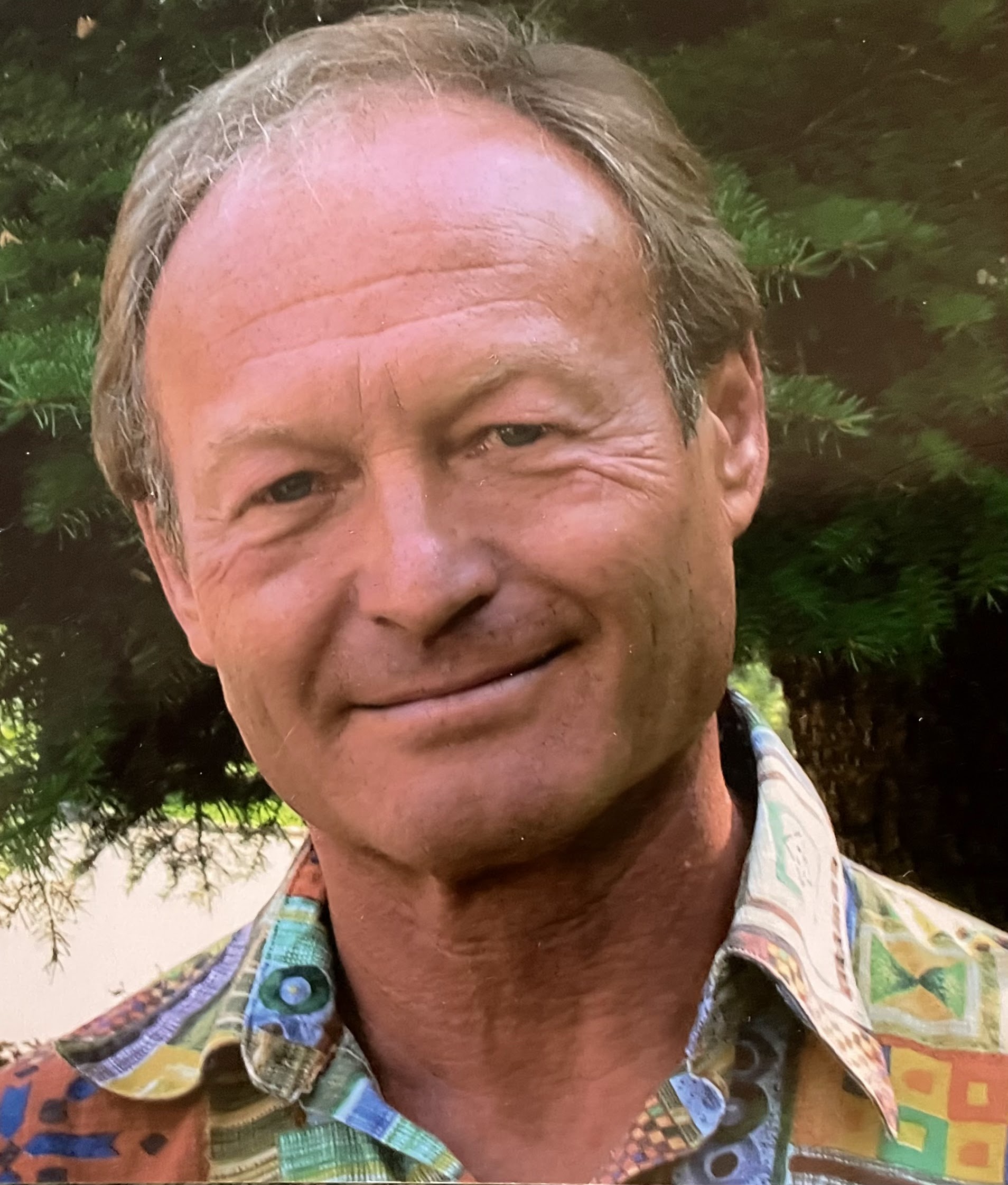 A photo of author Lloyd Ratzlaff is a light-skin-toned man with short sandy-blonde hair. He is standing in front of a tree and smiling into the camera.