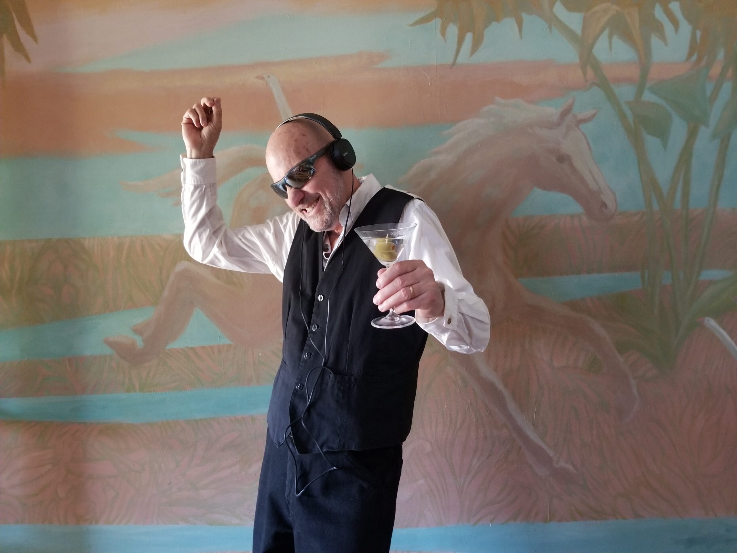 A photo of author Stan Rogal. He is a bald light skin-toned man wearing a black vest over a white-collared shirt and black dress pants. He is holding a dance pose with a martini in his hand, and wearing headphones. He is in front of a wall with a mural of white horse. 