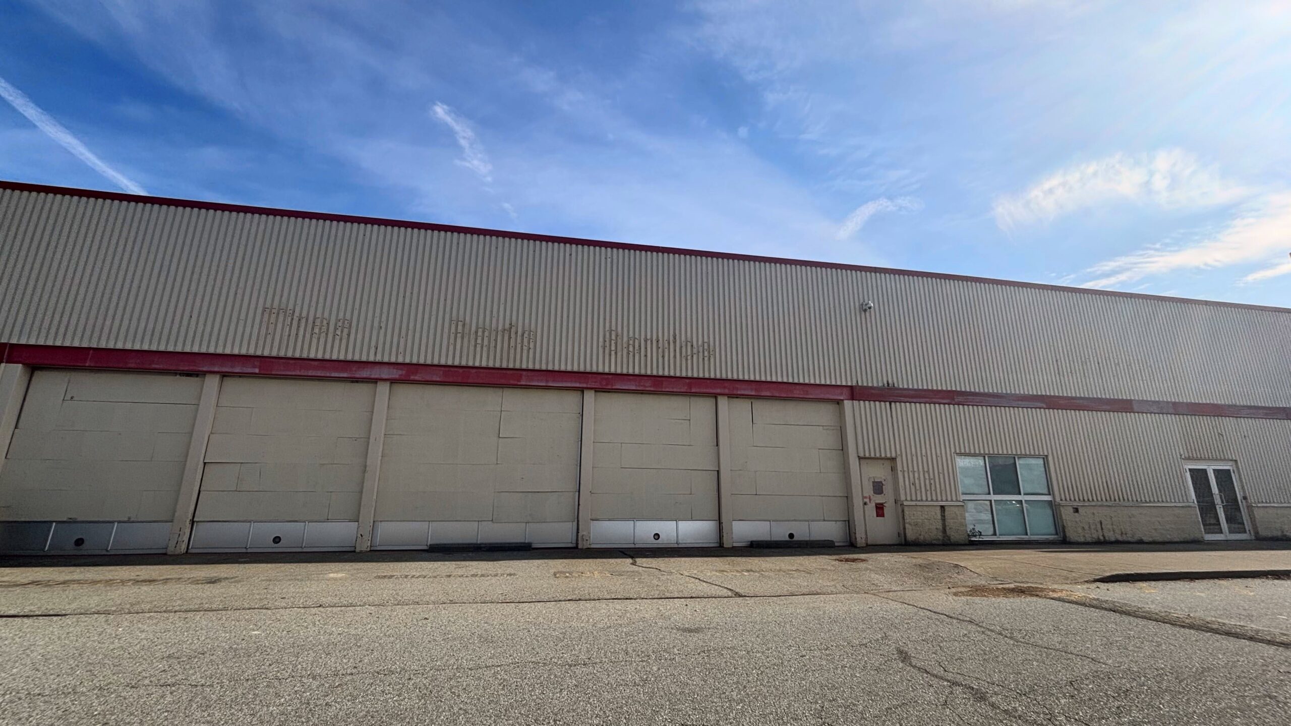A photograph of an abandoned car service building, the service bays are bricked up, and the ghosts of the "Tires, Parts, Service" signage are still visible on the walls. The sky is bright blue overhead.