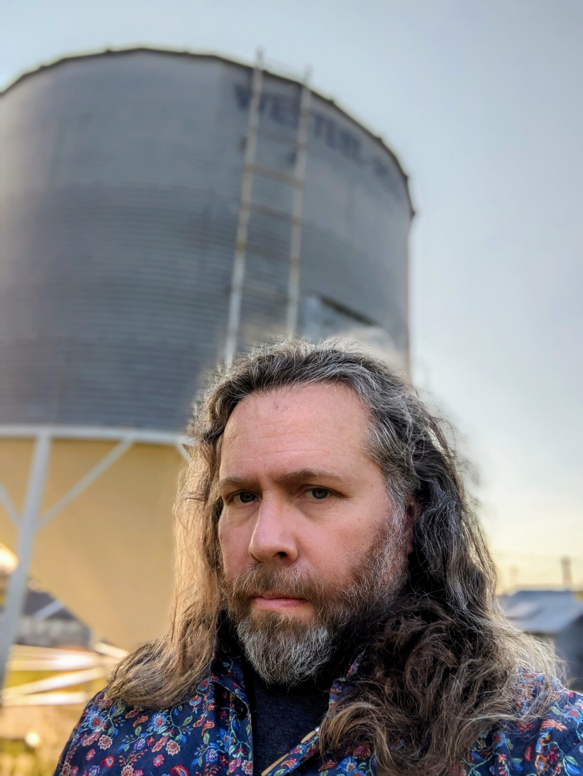 A photo of poet D.A. Lockhart. He is a light skin-toned Indigenous man with long, grey-brown hair and a grey-brown beard, wearing a floral paisley printed blue button down shirt. He stands in front of a water reservoir tower at dusk, the sky darkening behind him.