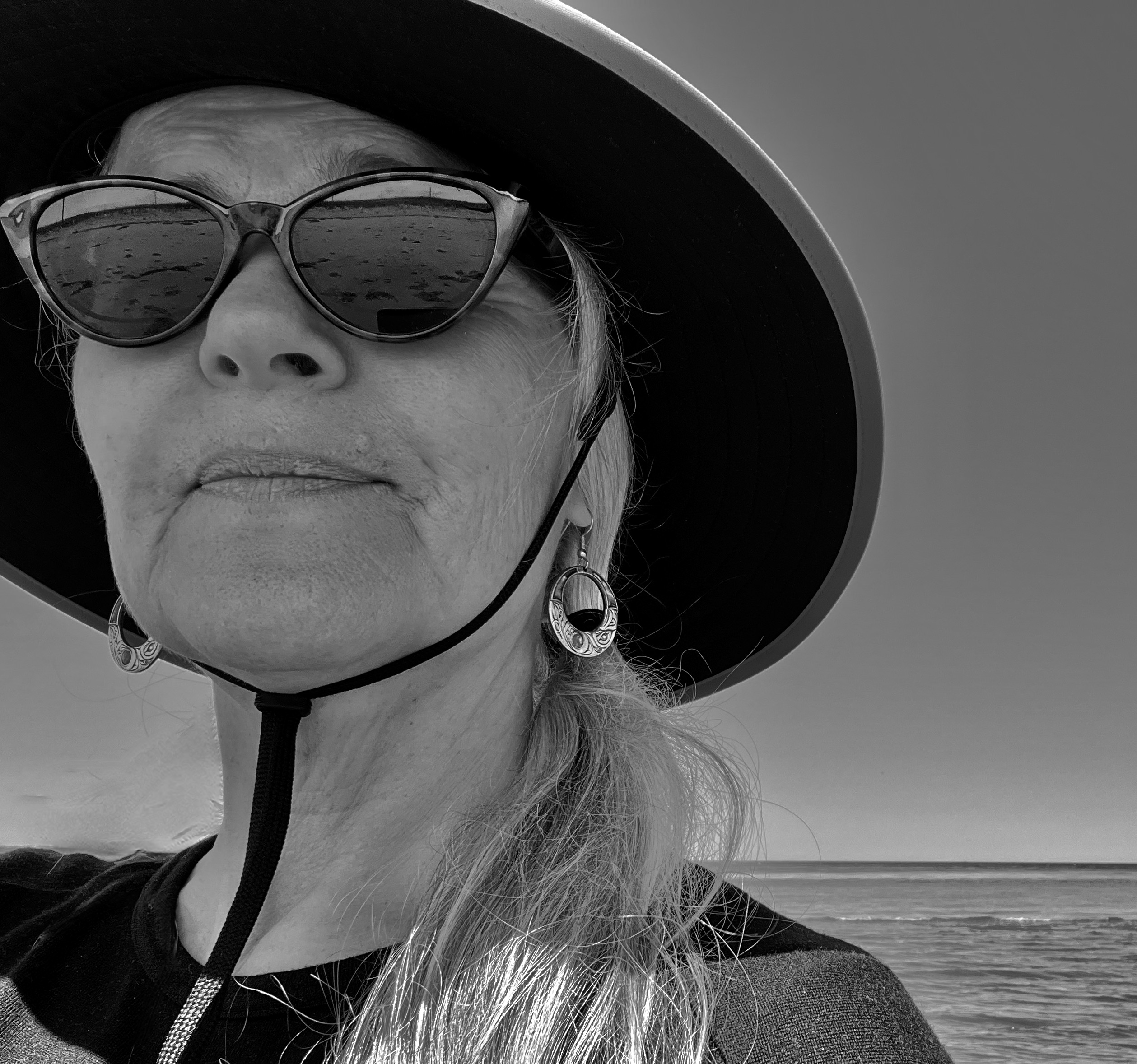 A black-and-white photo of author patti sinclair. She is a light-skin-toned woman with blonde hair. She is wearing a large-brimmed hat with sunglasses and is standing in front of a large body of water.