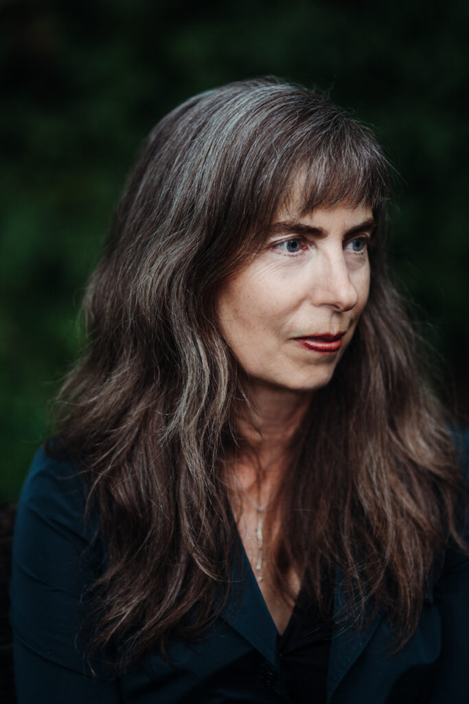 A photo of Catherine Bush. She is a light-skin-toned woman with long brown and greying hair. She is looking off into the distance. 