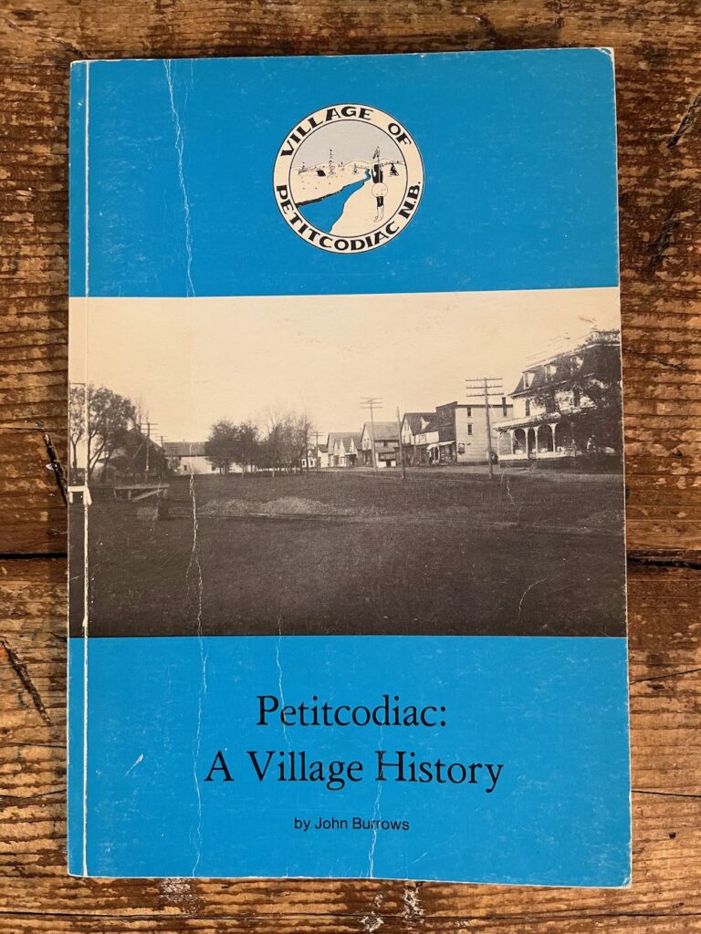 A copy of the book Petitcodiac: A Village History, by John Burrow. Blue horizontal bands surround an archival photograph of the main street of the town.