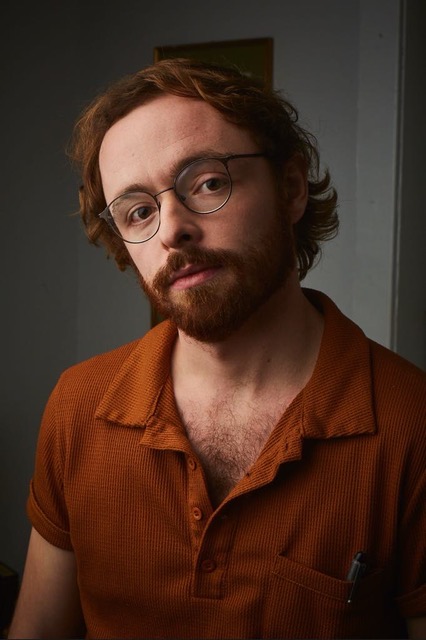 A photo of Curtis John McRae. He is a light-skin-toned man with red hair and a short beard. He is wearing an orange collared shirt and wire-rimmed glasses and looks into the camera. 