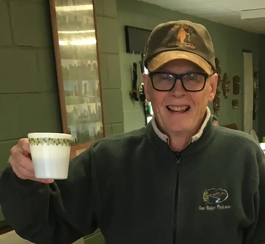 A photo of writer David Carpenter. He is an older, light skin-toned man with thick-framed black glasses, wearing a baseball hat and matching green jacket, toasting toward the camera with a white coffee mug.
