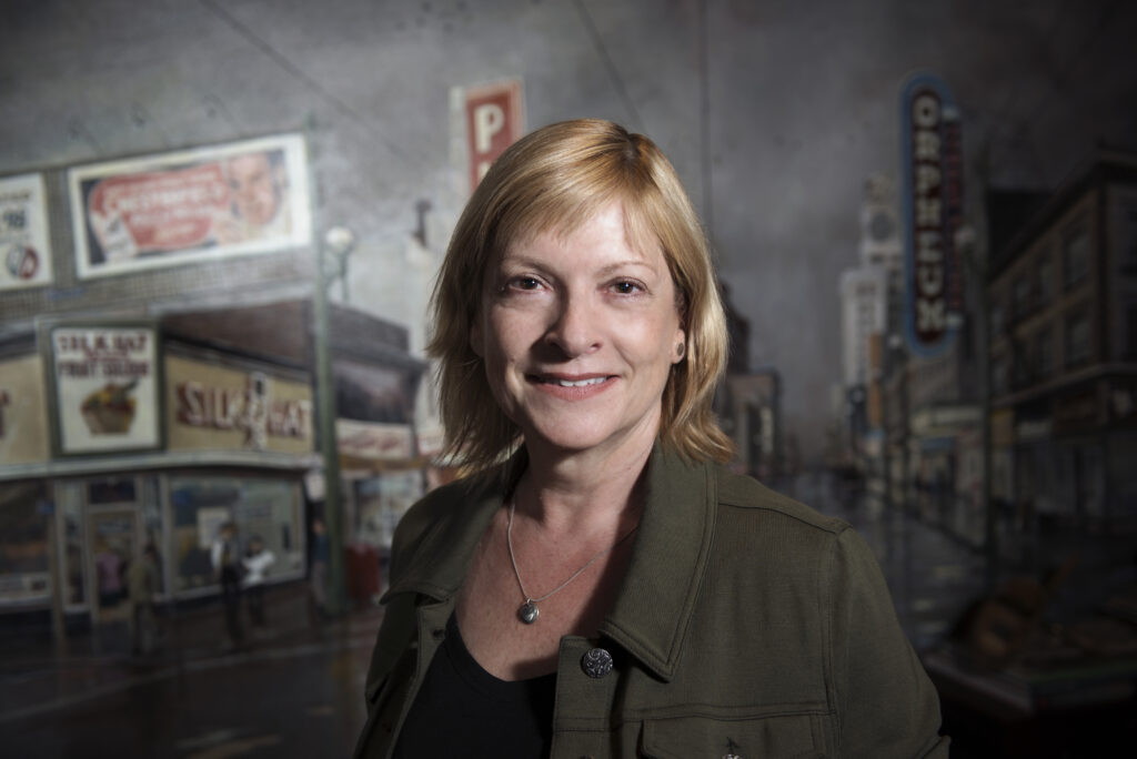 Photo of Eve Lazarus. She is a light-skin-toned woman with chin-length blonde hair. She is wearing a black shirt and a dark green jacket and smiling into the camera.
