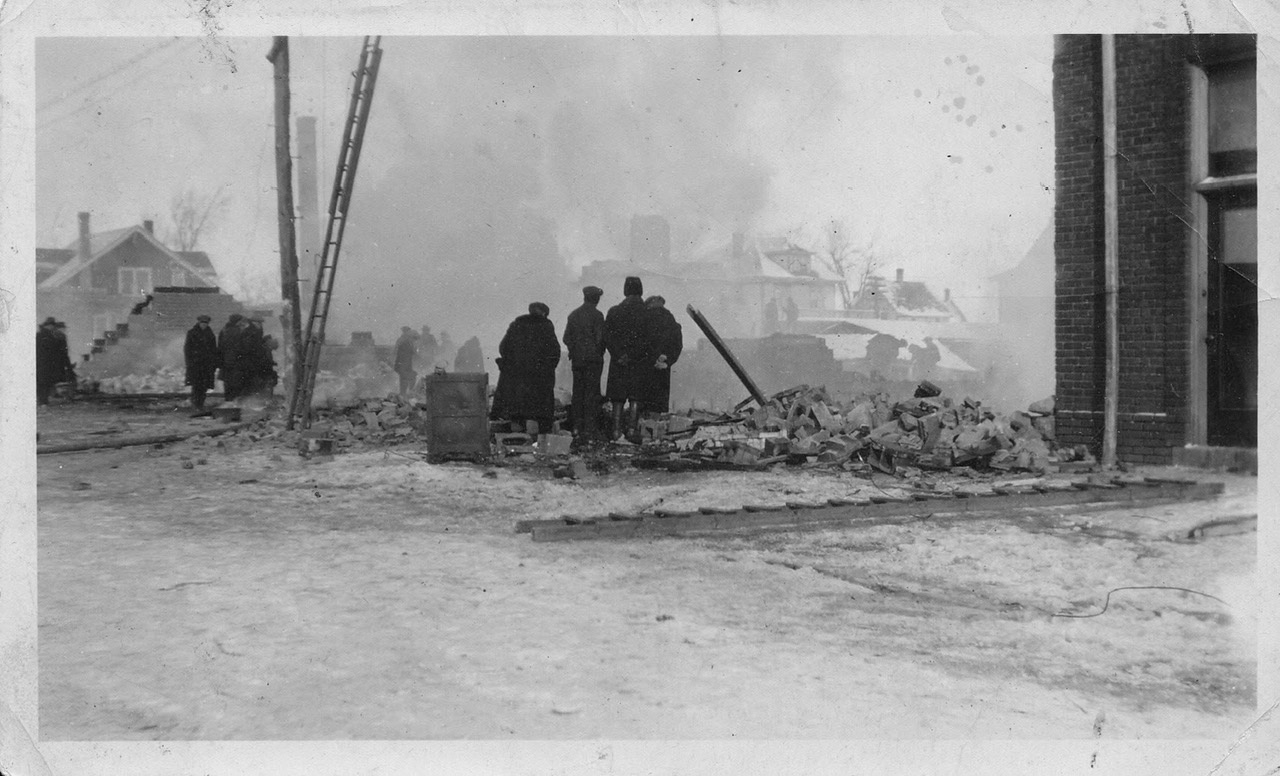 An archival photograph of one of the fires in Petitcodiac, showing several figures silhouetted against smoke and rubble.
