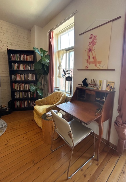 A photo of Curtis John McRae's writing space. There is a bookshelf in one corner with a large plant beside it. A mustard-yellow suede armchair sits in front of a large window and a small wooden desk and chair are beside it.