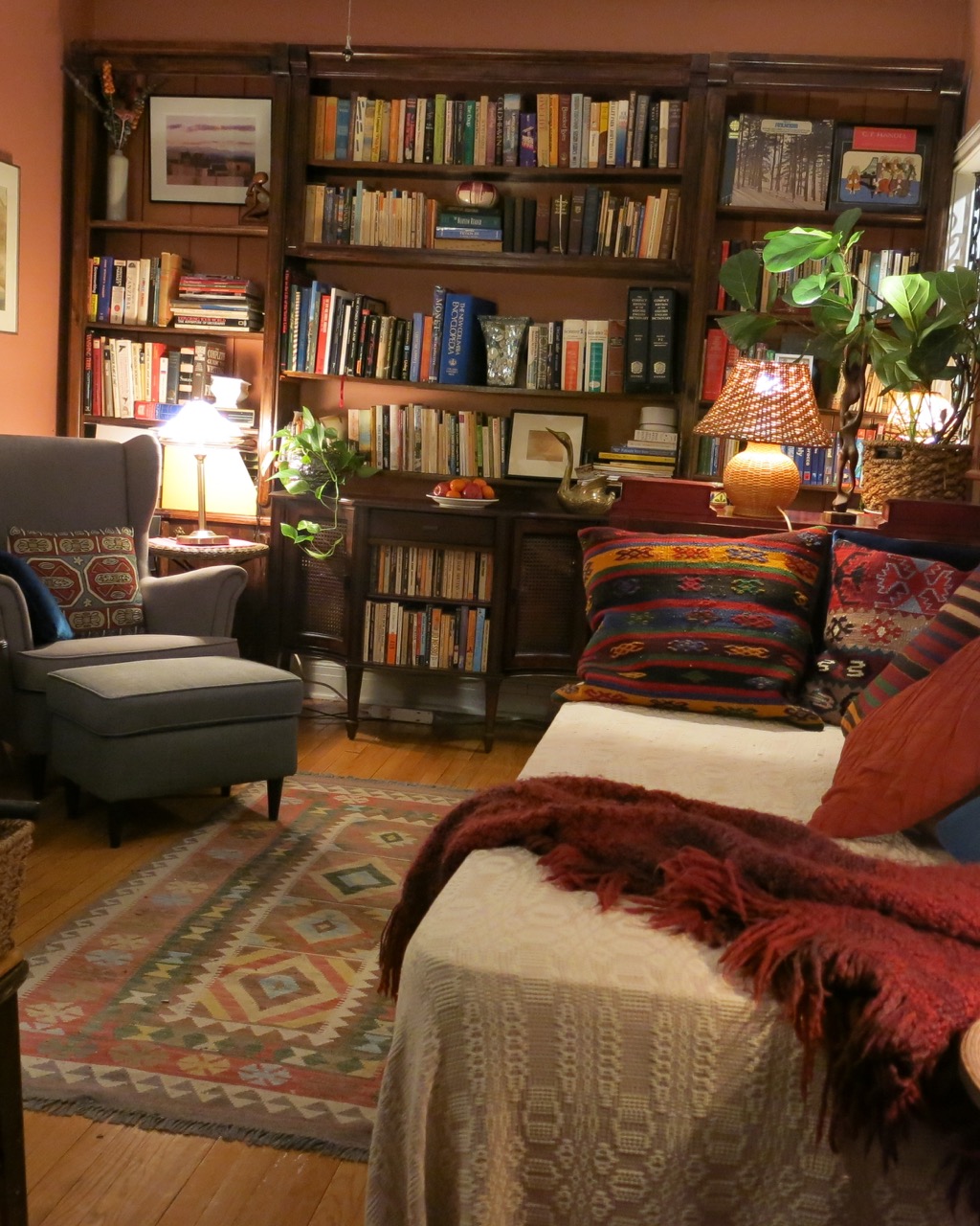 A photo of Irena Karafilly's study. It is painted in a warm coral colour, with lots of rich textiles in the rug, throw pillows and blankets. A built-in dark wood bookcase lines the back wall, stocked full with books. All of the table lamps are on, bathing the room in a warm light.