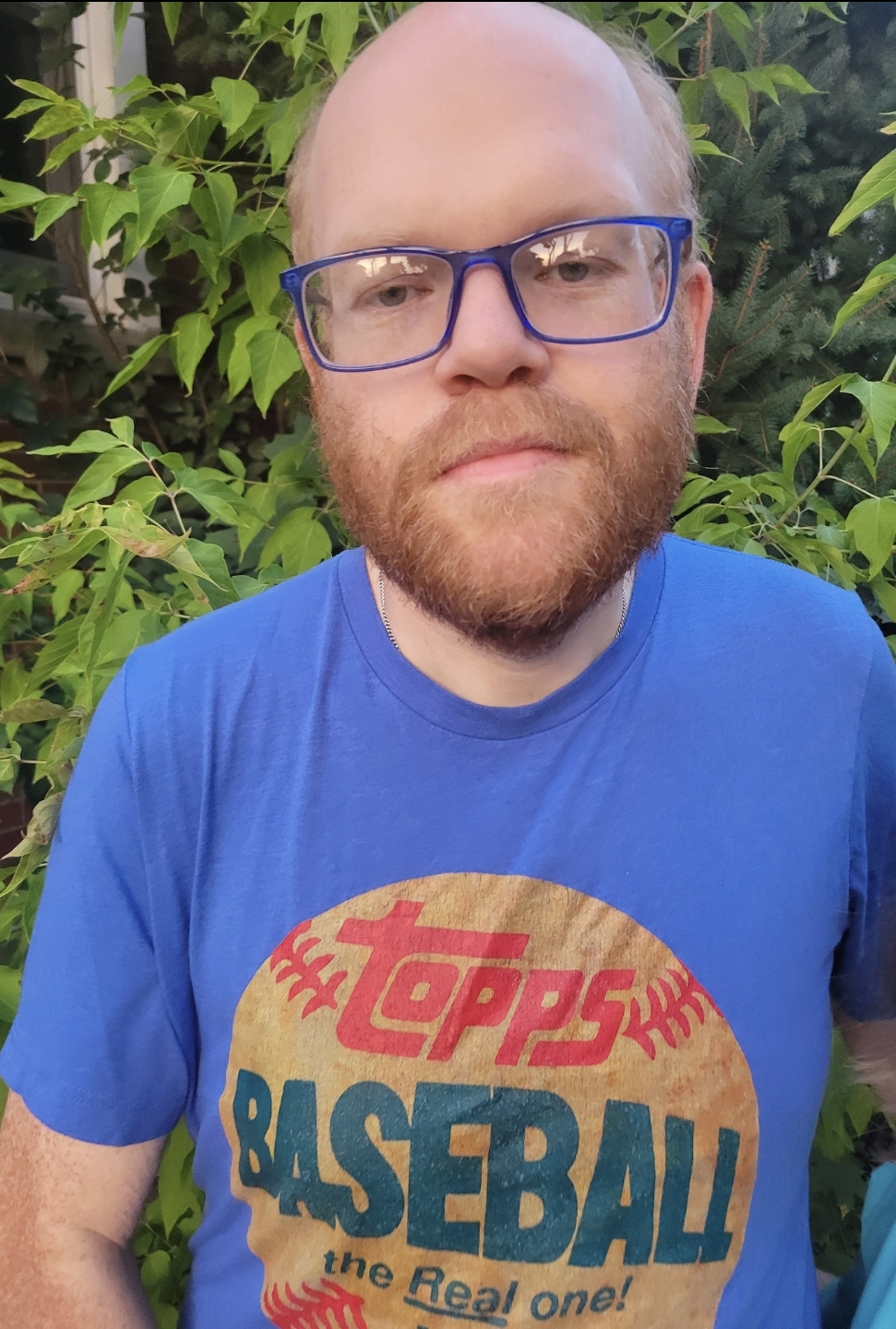 A photo of Jacob McArthur Mooney. He is a light-skin-toned man with facial hair and large blue-rimmed glasses. He is wearing a blue t-shirt with writing on it that reads "Topps Baseball, the Real One." He is outside in front of a leafy hedge. Photo credit is Alexis von Konigslow.