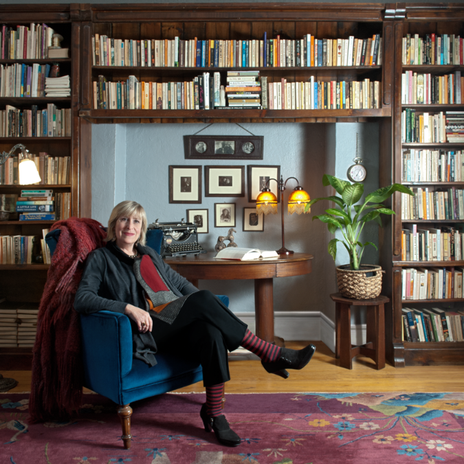 A photo of writer Irena Karafilly in a book-lined study. She is a light skin-toned woman with grey-blonde hair cut into bangs, sitting at ease in a navy armchair with her legs crossed.