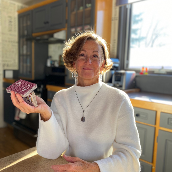 A photo of writer Lee Kvern. She is a light skin toned woman with short brown hair, wearing a white long sleeved shirt. She sits in a bright kitchen at the table, smiling, holding a deck of cards cut in two in her aloft hand.