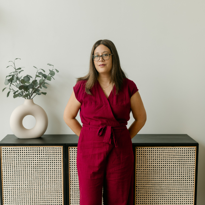 A photo of writer Teri Vlassopoulos. She is a medium skin toned woman with long brown hair, wearing glasses and a deep red linen jumpsuit. She stands in a minimally-decorated room, leaning against a credenza, with eucalyptus branches in a vase behind her right side.