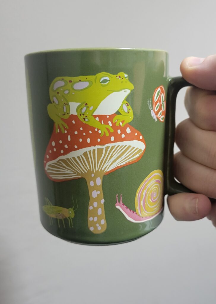 A photo of Jacob McArthur Mooney's coffee mug. It is a green mug with a drawing of a toad on top of a mushroom. There is a snail and other insects around the mushroom. 