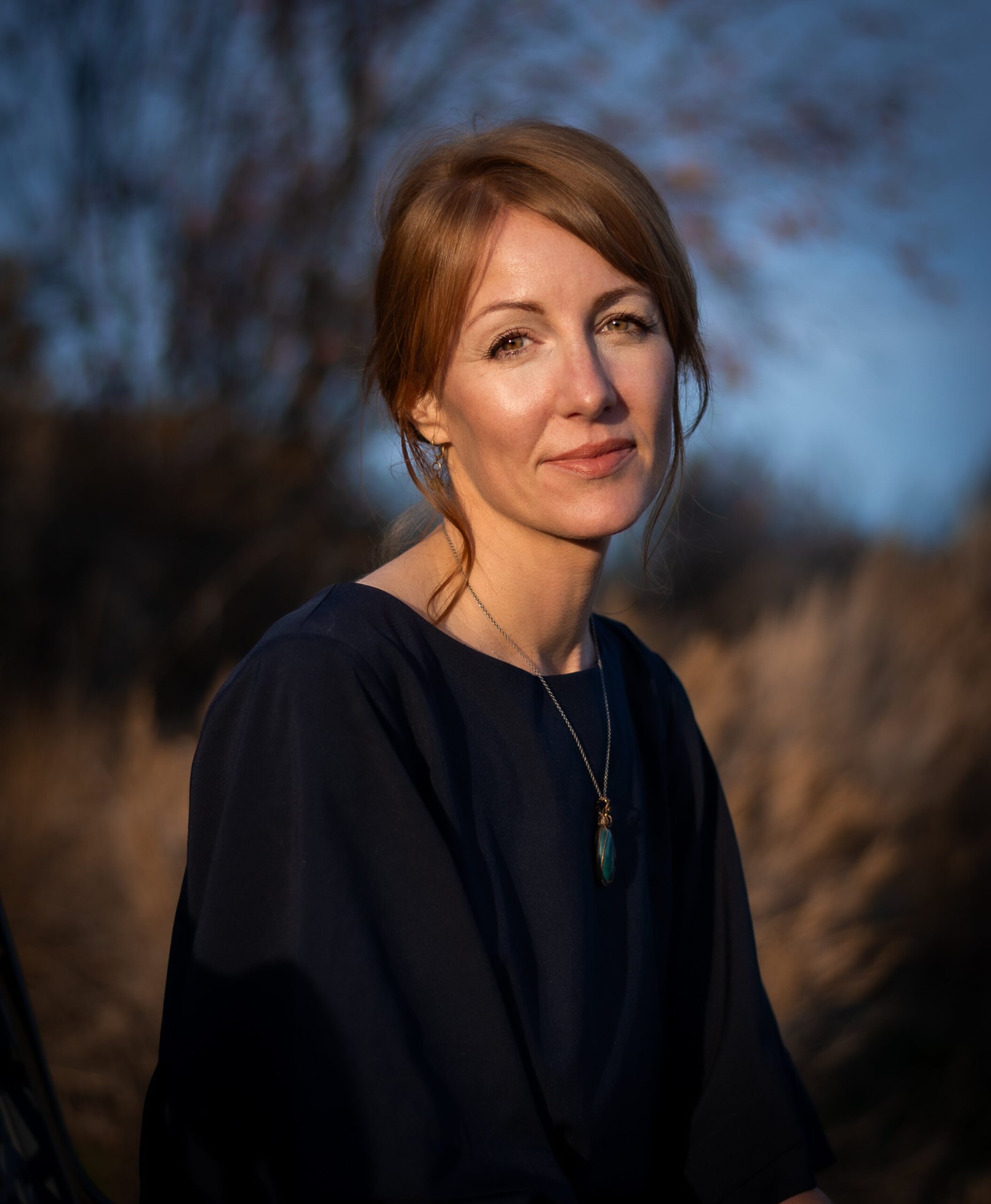 A photo of writer Natalie Appleton. She is a light skin toned woman with auburn coloured hair pulled back, wearing a dark sweater and a stone pendant. She sits outside, with wild grasses and a bare tree in soft focus behind her.