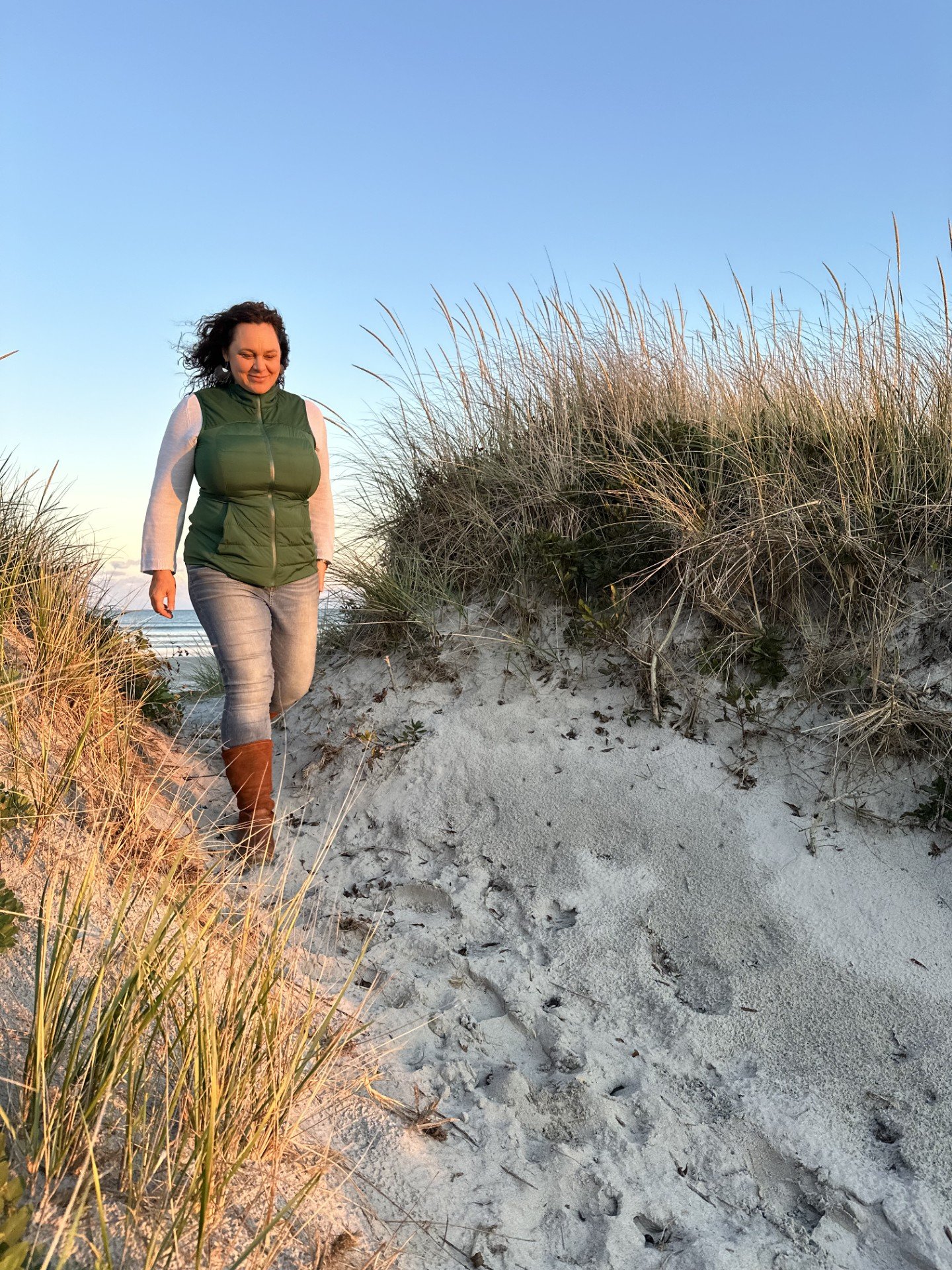 A photo of poet Jessica Hiemstra walking on a beach. She is a medium skin-toned woman with curly dark hair, wearing a zip up puffy green vest over a light long-sleeved shirt and jeans. She walks between two sand dunes covered in tall grasses, on a well-trod path. The water is behind her, and the wind is whipping through her hair.