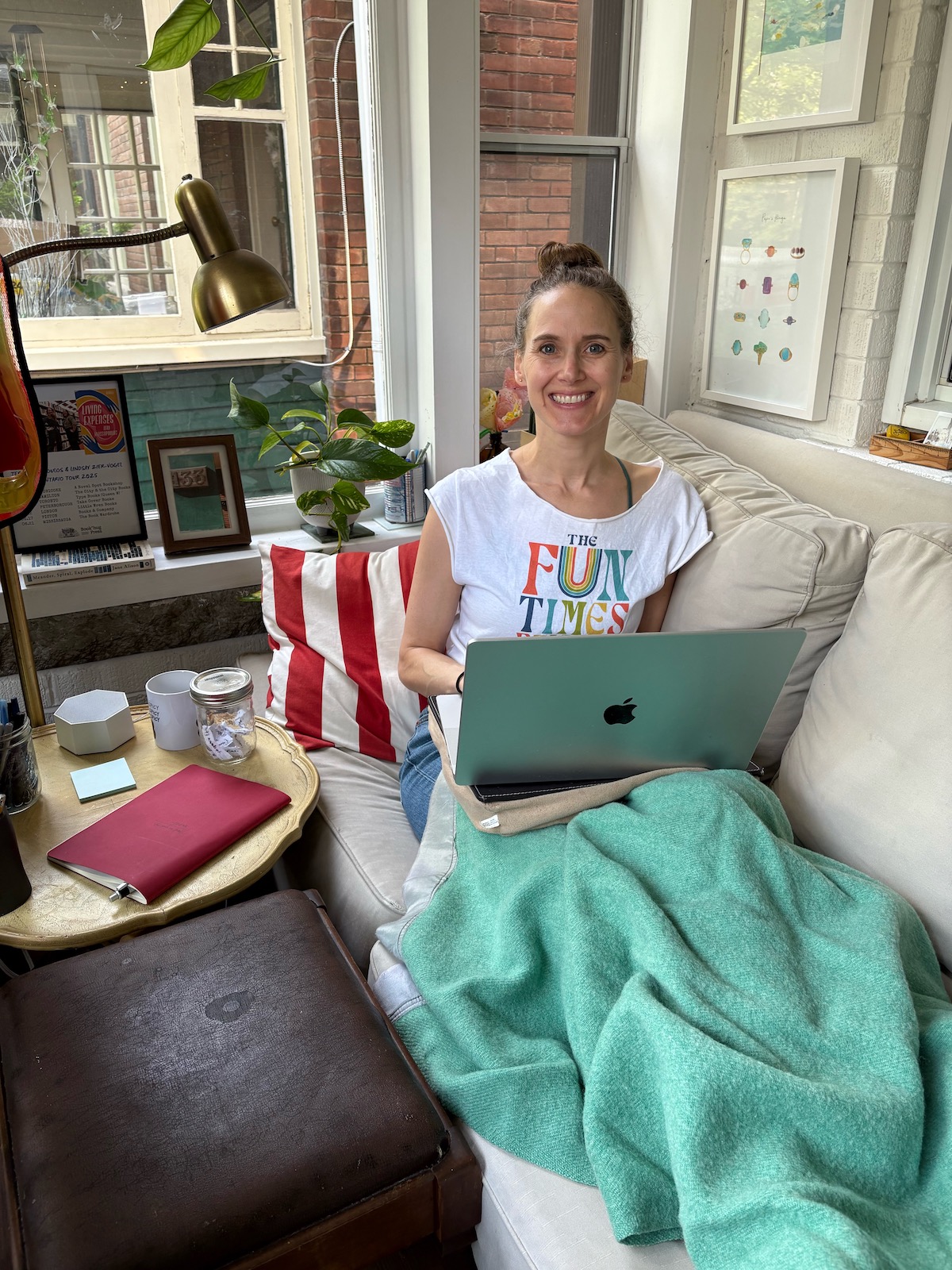 A photo of Lindsay Zier-Vogel in her front sun room. She is sitting on the sofa and smiling, a blanket over her legs and a laptop in her lap. She wears a Fun Times Brigade t-shirt. There is art on the walls and a tray with a notebook and mug next to her. The room is bright, busy, and inviting.